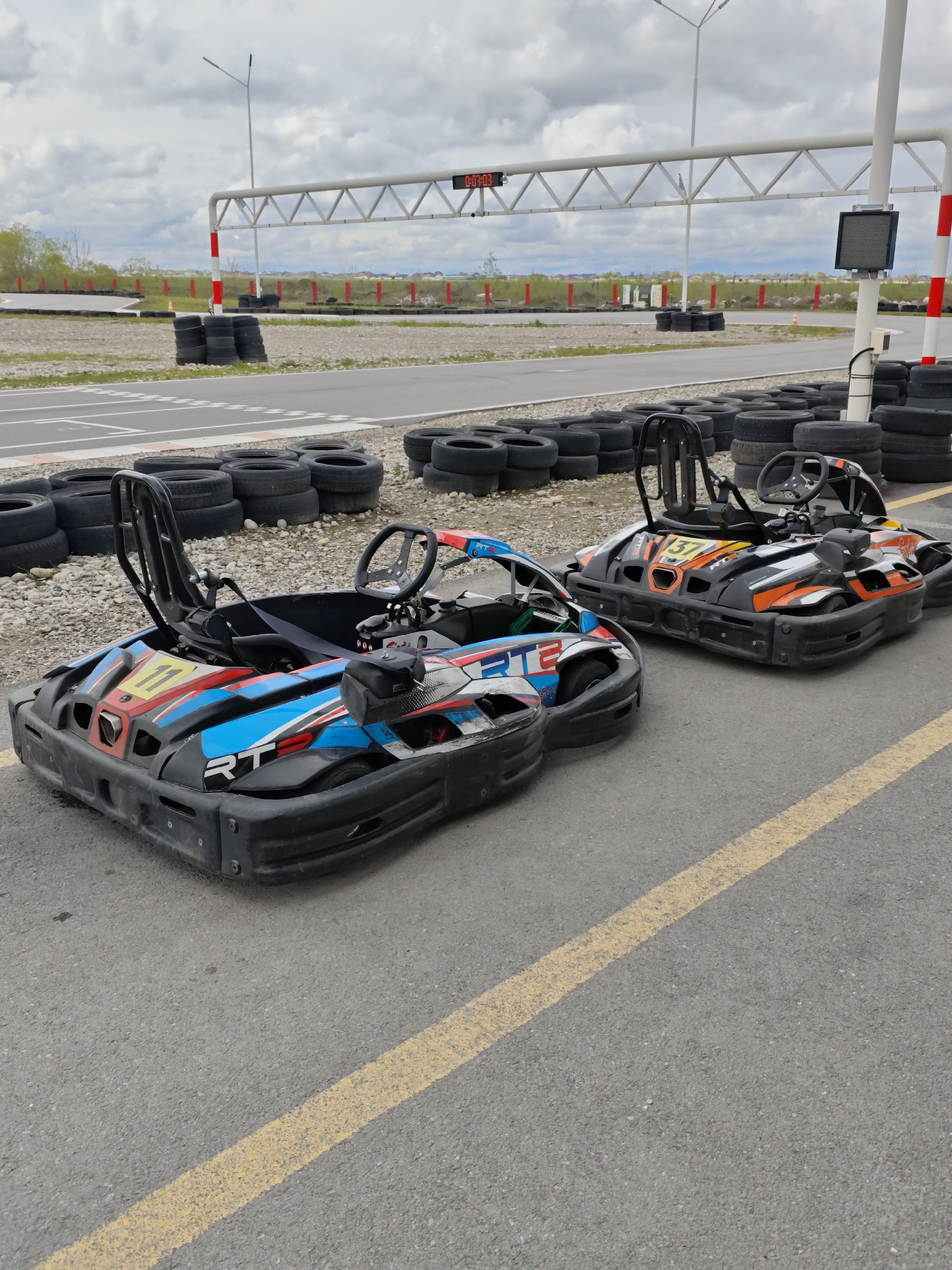 The image depicts two go-karts parked on an asphalt surface marked with yellow lines, likely at a go-kart racing facility. The go-karts, one predominantly blue and the other orange, are facing towards the background, where a white metal gantry structure with a digital timer is visible. Stacks of black tires are arranged around the perimeter of the track and adjacent to a pebbled area. The backdrop shows a distant landscape under an overcast sky. The city and country are stated as Kishpek, Russia.