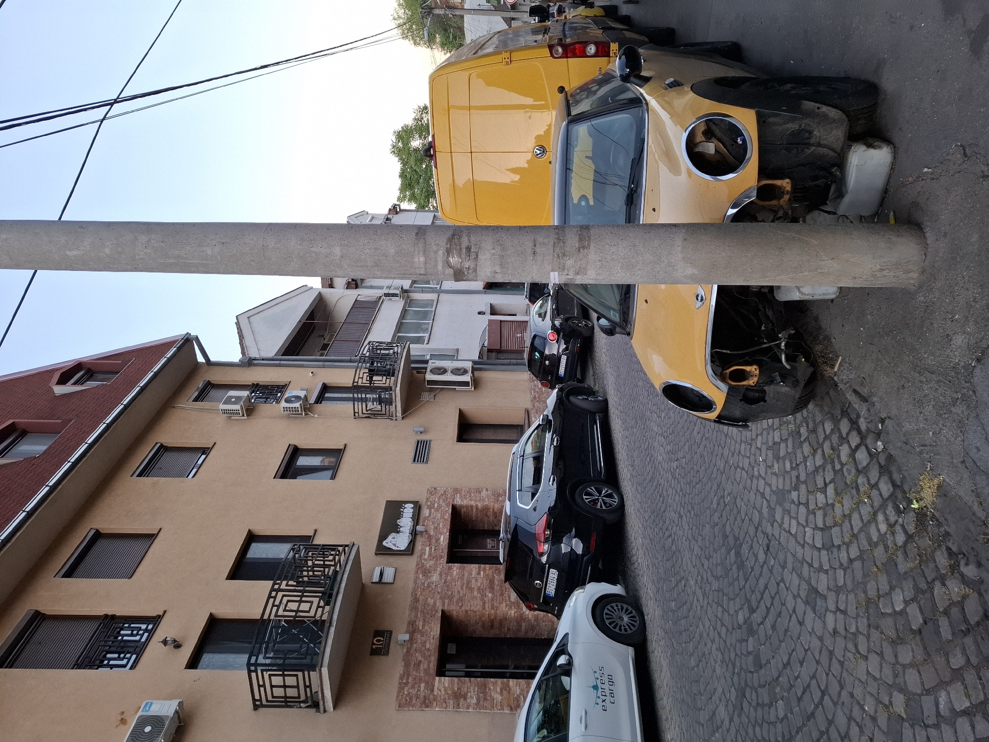 The image depicts a street scene in Zemun Urban Municipality, Serbia. A yellow Volkswagen van and a damaged yellow Mini Cooper are parked along the side of the street. Behind them, a black SUV and a white "Express Cargo" vehicle are also parked. The street is paved with cobblestones. Adjacent to the parked vehicles is a building with beige facade, adorned with balconies and air conditioning units. Street lights and overhead wires are also visible.