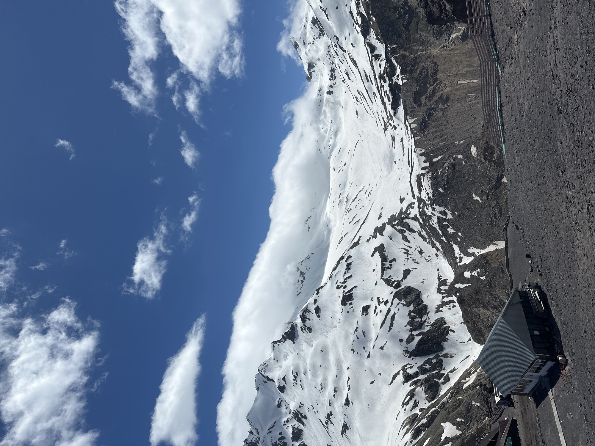 The image shows a view of snow-capped mountains with some dark, rocky slopes. In the foreground, there is a dark, gravelly ground and a long, dark-colored building with a grey roof and white walls. The building appears to be a structure like a shelter or lodge. Further back, there are more buildings visible on the left side of the frame. A wooden fence runs along the middle ground, separating the foreground from the mountainous terrain. The sky is a vibrant blue with scattered white clouds. The scene suggests a location in a mountainous, possibly cold climate, consistent with the city of Terskol, Russia, which is known for its proximity to Mount Elbrus, a major Caucasian peak.