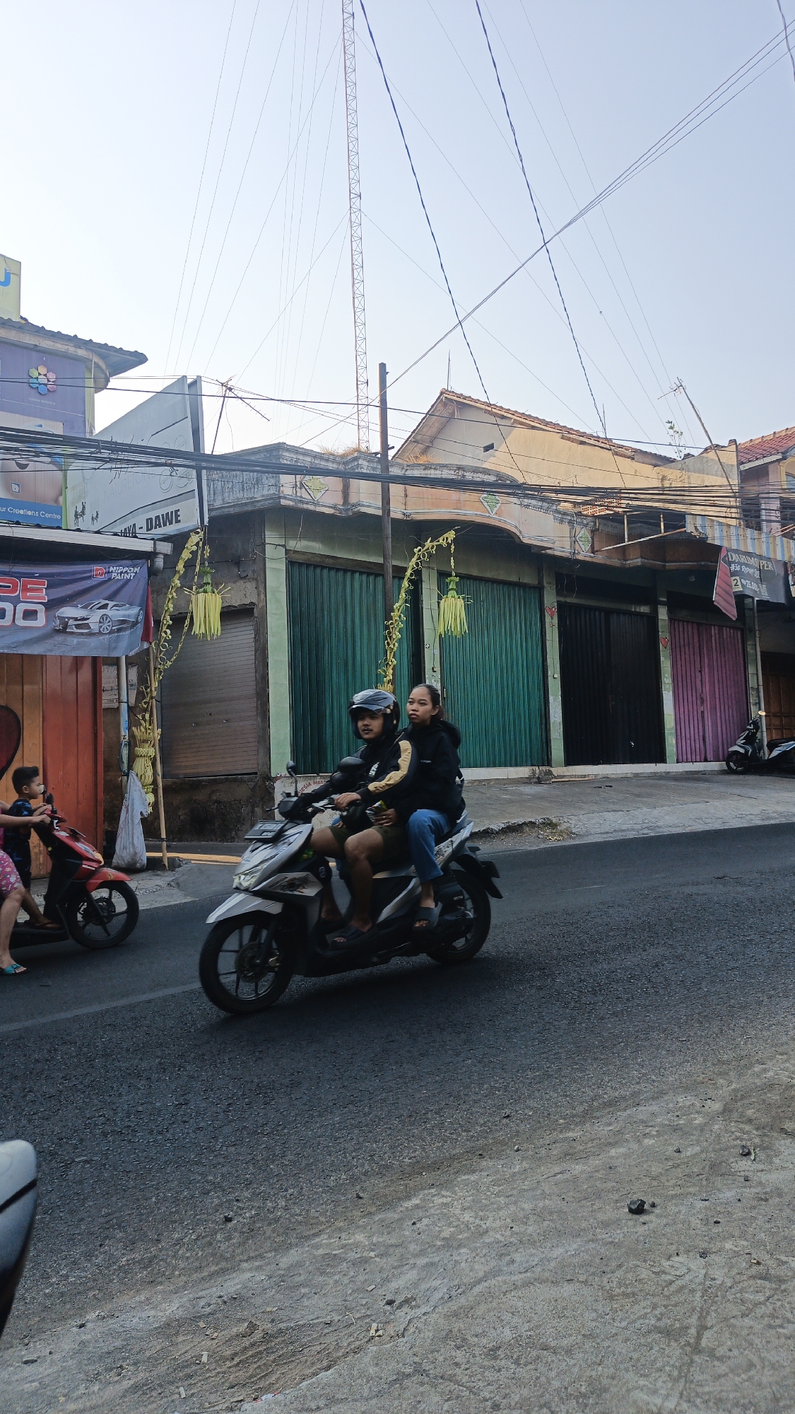 The image depicts a street scene in Dawe, Indonesia. In the foreground, a motorcycle carrying two individuals is traveling on the asphalt road. The driver, a male wearing a helmet, is operating the vehicle, while a female passenger sits behind him. They are both dressed in casual clothing. To the left of the main motorcycle, another motorcycle with a young child and an adult are visible. The background features a row of buildings with shops, characterized by colorful shutters and awnings. Overhead, numerous electrical wires crisscross the sky, with a tall transmission tower visible in the distance. The lighting suggests it is daytime, with the sun casting shadows. The environment appears to be an urban or semi-urban area in Indonesia.