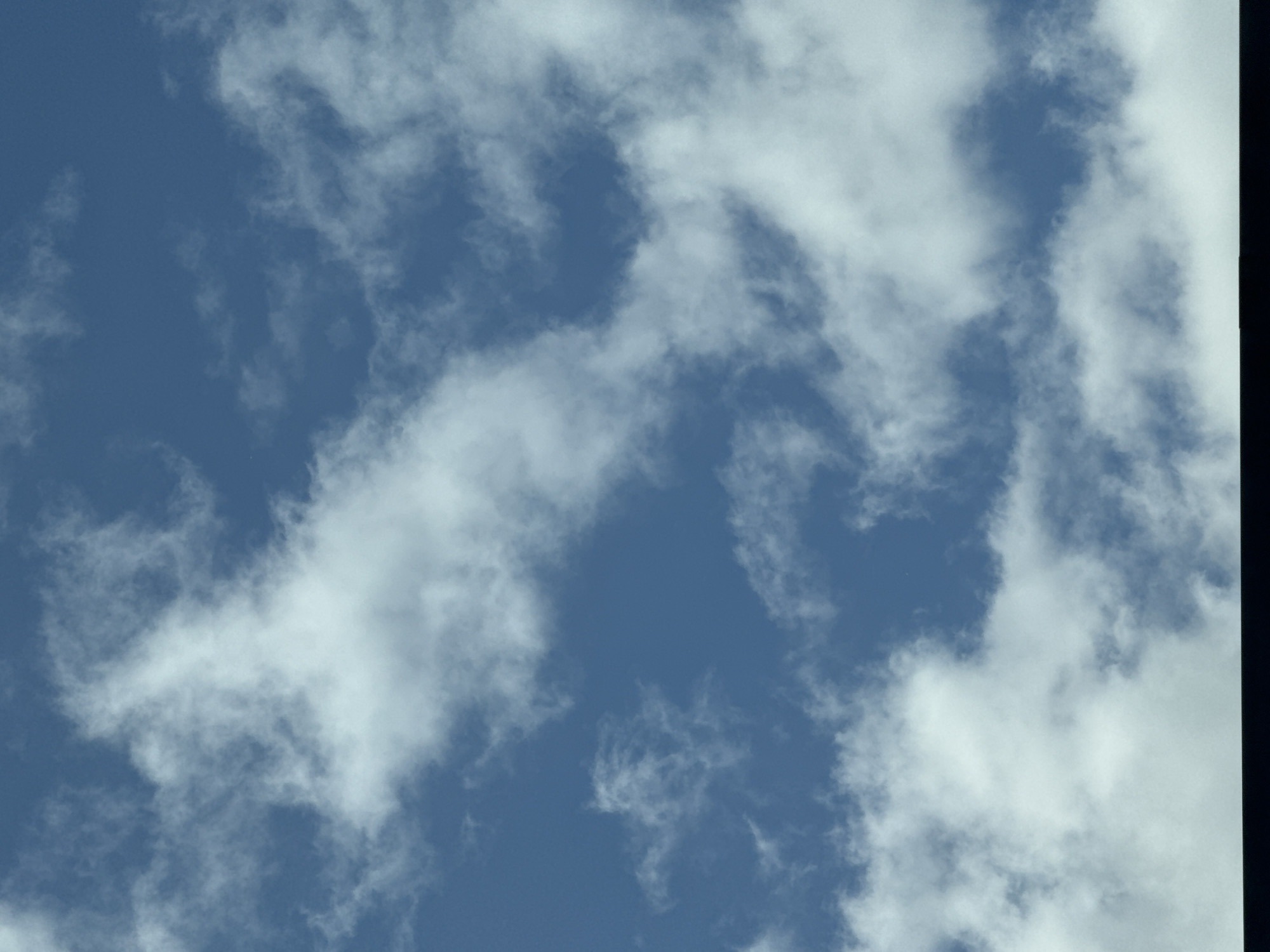 This is a vertical, full-frame photograph of a blue sky with scattered white clouds. The clouds vary in density and shape, with some appearing wispy and others more substantial and opaque. The lighting suggests it is daytime, likely mid-morning or afternoon, with the sun not directly visible but illuminating the clouds from above. There is no indication of any urban environment or specific landmarks within the image itself. Without additional context or visual cues, it is impossible to determine if this image was taken in Waterford, Ireland, or any other specific location. The image is purely of the sky.