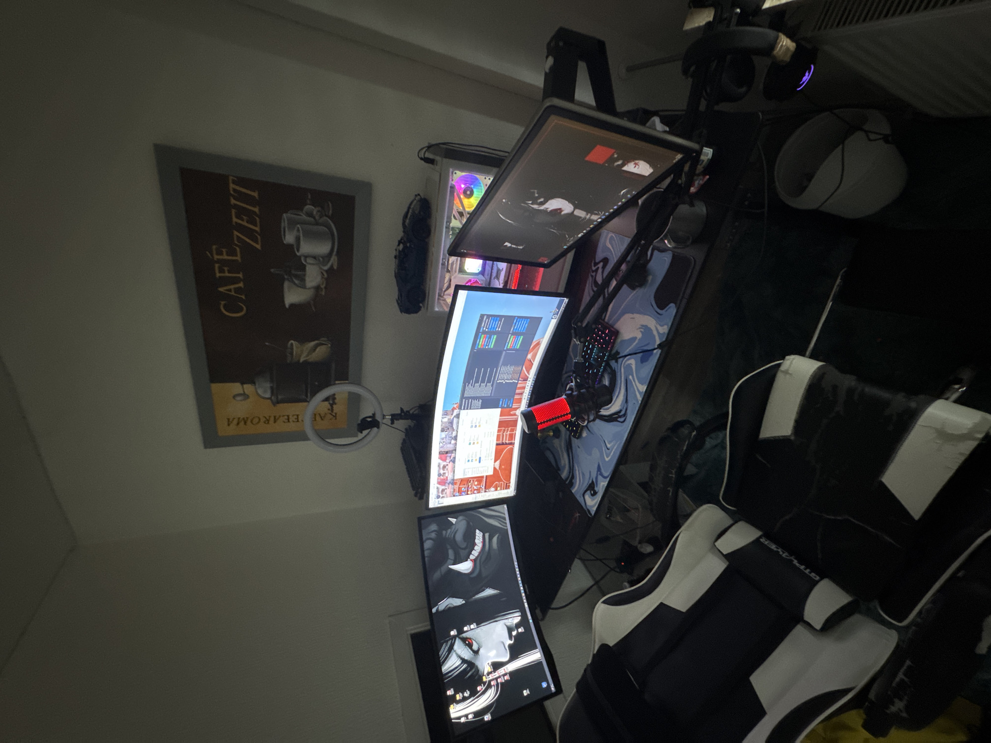 This media file depicts a gaming setup in a room, likely in Neubrandenburg, Germany. The scene is oriented sideways, showing a desk with multiple computer monitors arranged vertically and horizontally. The top monitor displays a dark image with red and white elements. Below it, another monitor shows a complex interface with various colored bars. The bottom-most monitor is positioned vertically and displays an anime-style image of a woman with red eyes and dark hair.
In the background, a PC tower with a glowing, rainbow-colored fan is visible. A ring light is mounted above the setup, and a microphone with a red pop filter is attached to an arm. Headphones are seen resting on top of the PC monitor mount.
To the left of the monitors, a framed poster with the text "CAFÉ ZEIT" and images of coffee cups is hanging on a light-colored wall. A ring light is also visible near the poster.
The foreground is dominated by a black and white gaming chair, identified by the text "GTPLAYER" on its headrest. The chair is partially obscured by darkness, but its distinctive design is clear.
The room appears to be dimly lit, suggesting it might be evening or night. The overall impression is that of a functional and personalized gaming or workstation.