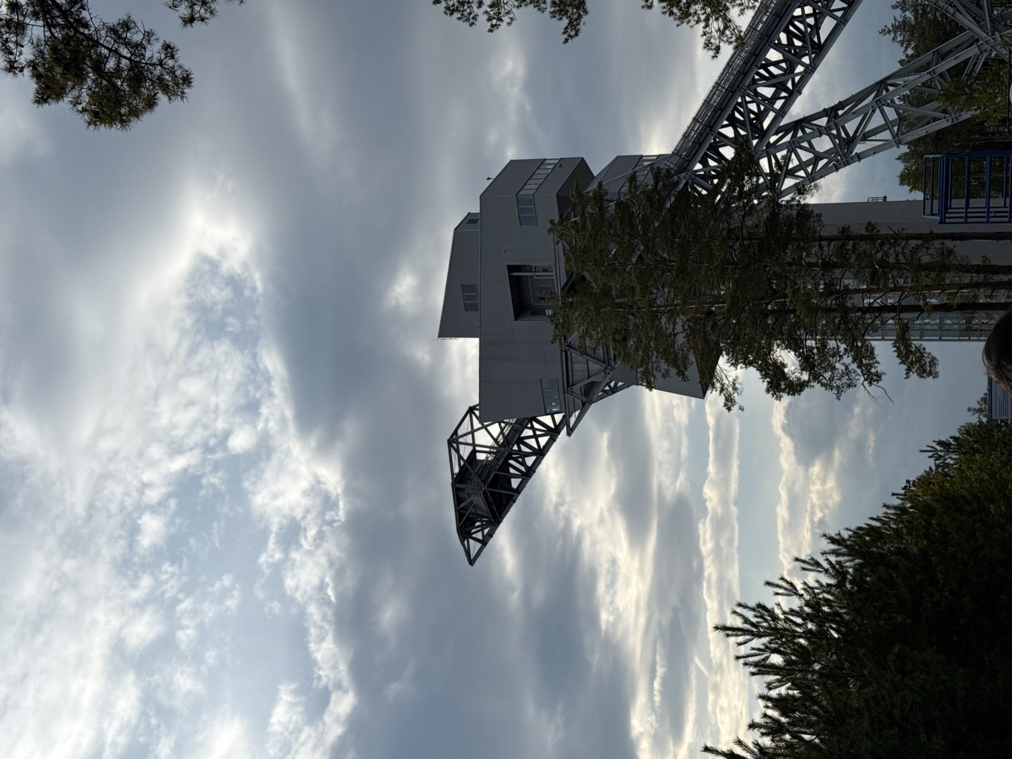A low-angle view of a tall structure in Прикамский, Russia, likely an industrial facility or observation tower. The structure features a combination of solid, angular architecture and exposed metal framework. The sky is overcast, with varied cloud formations. Trees are positioned along the frame's edges, some obscuring parts of the structure. The structure extends towards the top of the frame, set against the cloudy sky.