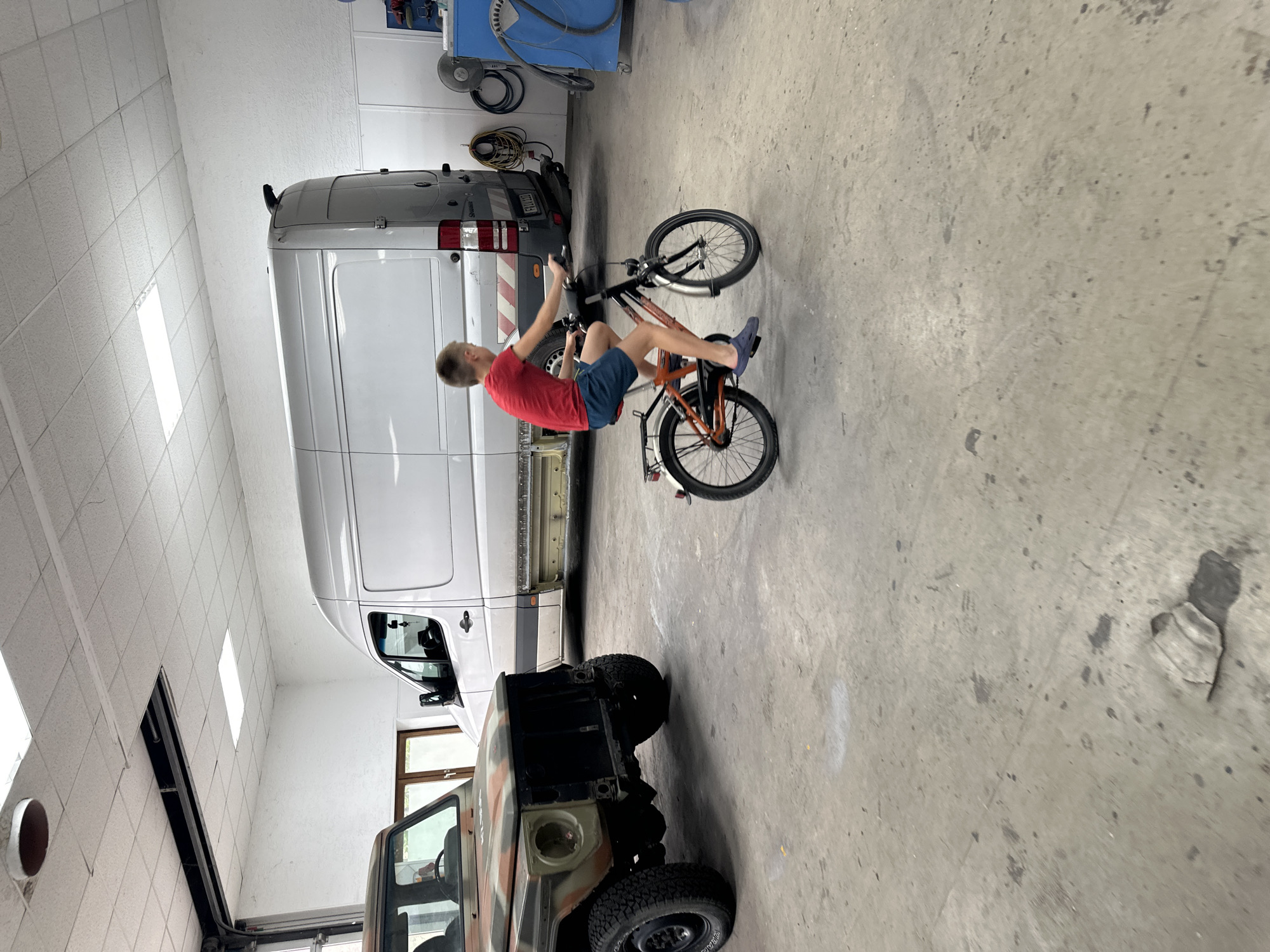 A young boy rides a bicycle inside a workshop or garage. The boy, wearing a red t-shirt and blue shorts, is positioned in the center of the frame, facing right. He is on an orange and black bicycle with black wheels and handlebars. In the background, a large, white van is parked to the right of the boy, and a camouflaged off-road vehicle is parked to the left. The floor is concrete, with various stains and markings. The walls are white, and the ceiling is made of suspended ceiling tiles with fluorescent lighting. The overall environment suggests a repair shop or storage area. The image was taken in Mainz, Germany.