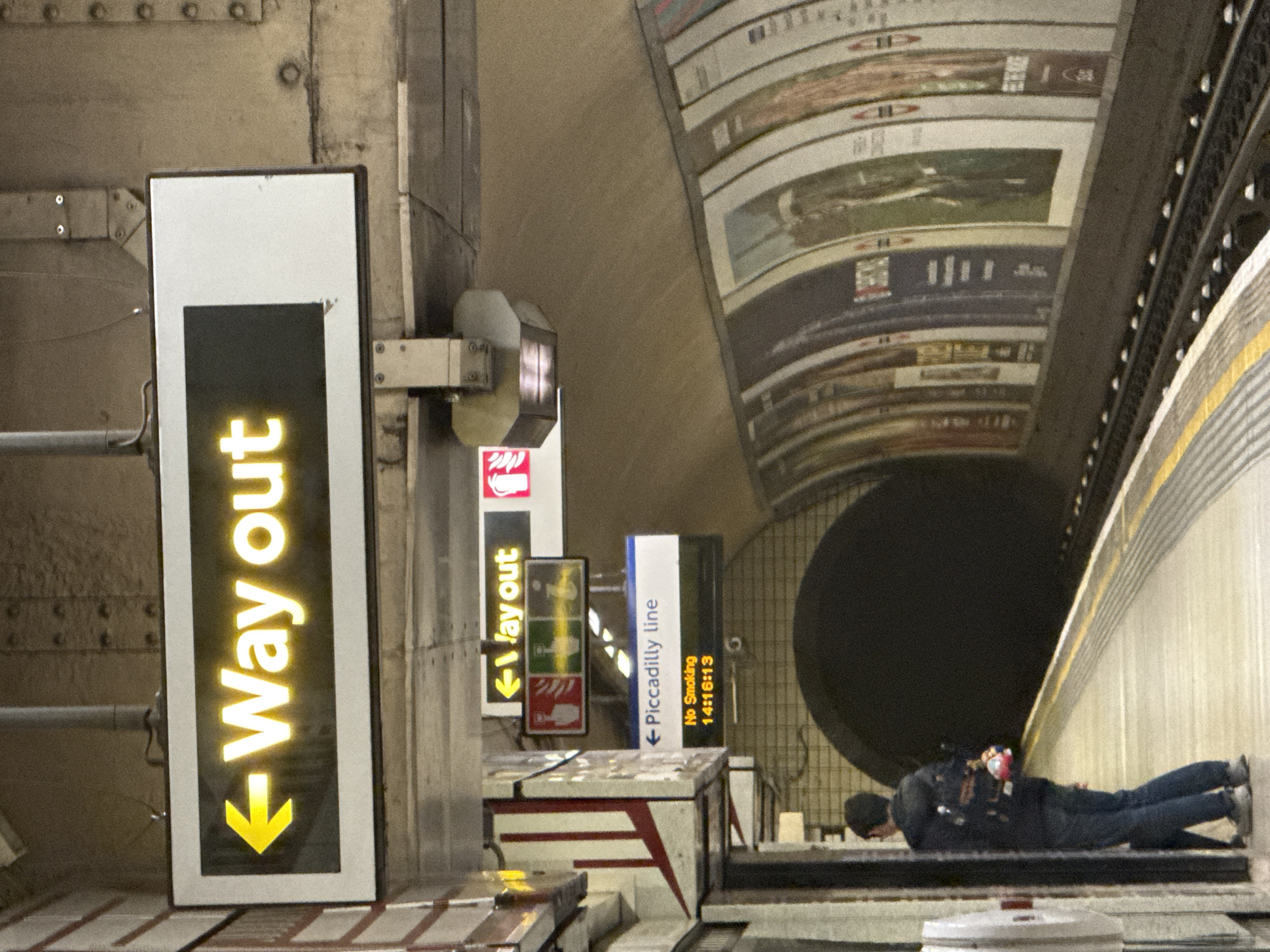 The image depicts a scene within a London Underground station. A man is lying prone on a platform, facing towards the tunnel. He is wearing dark clothing and a hat. To the left are several signs, including a prominent "Way Out" sign with a yellow arrow, a fire safety sign, and a sign indicating the Piccadilly Line. The station walls are lined with advertisements and there's overhead structural support. The scene suggests a potentially unsafe situation, with a person in close proximity to the tracks.