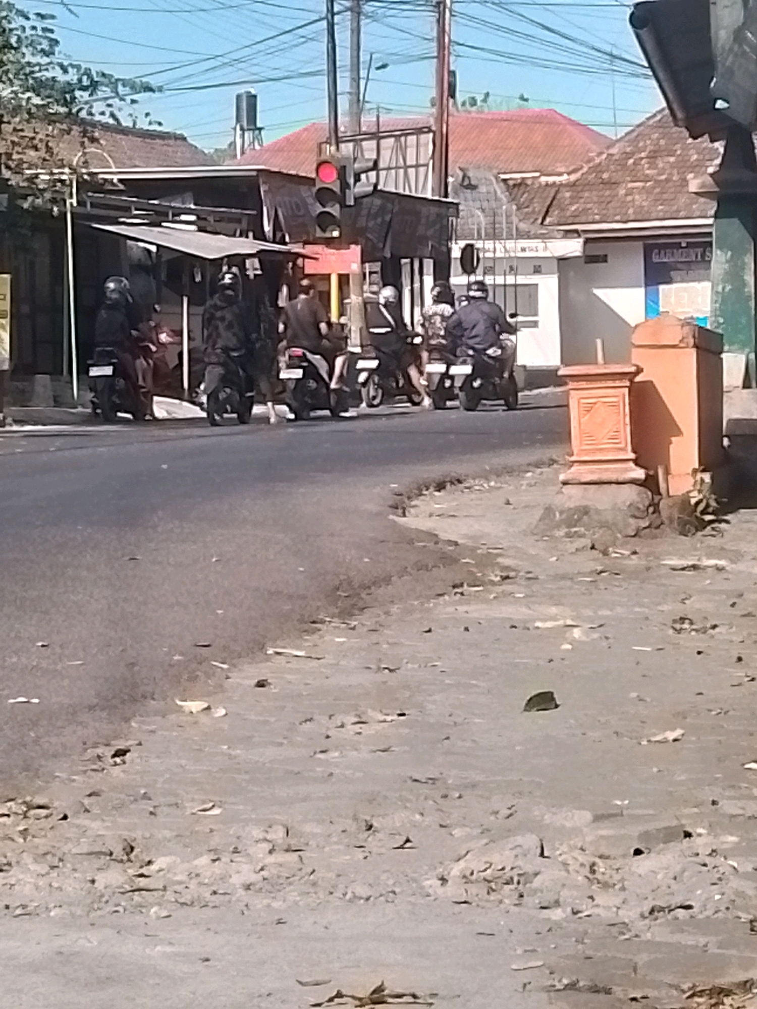 This is a street-level, eye-level, medium shot photograph taken in the city of Wonosari, Indonesia, on a sunny day. The image captures a group of people riding motorcycles on a paved road. The motorcycles are mostly dark in color, and the riders are wearing helmets. In the background, there are buildings with tiled roofs, typical of Indonesian architecture. A traffic light is visible, showing a red signal, suggesting the motorcycles are stopped or slowing down. The foreground of the image shows a rough, unpaved area with debris, likely the side of the road or a sidewalk. Power lines are visible crisscrossing the sky. The overall scene depicts a common urban environment in Indonesia, with motorcycles being a prevalent mode of transportation.