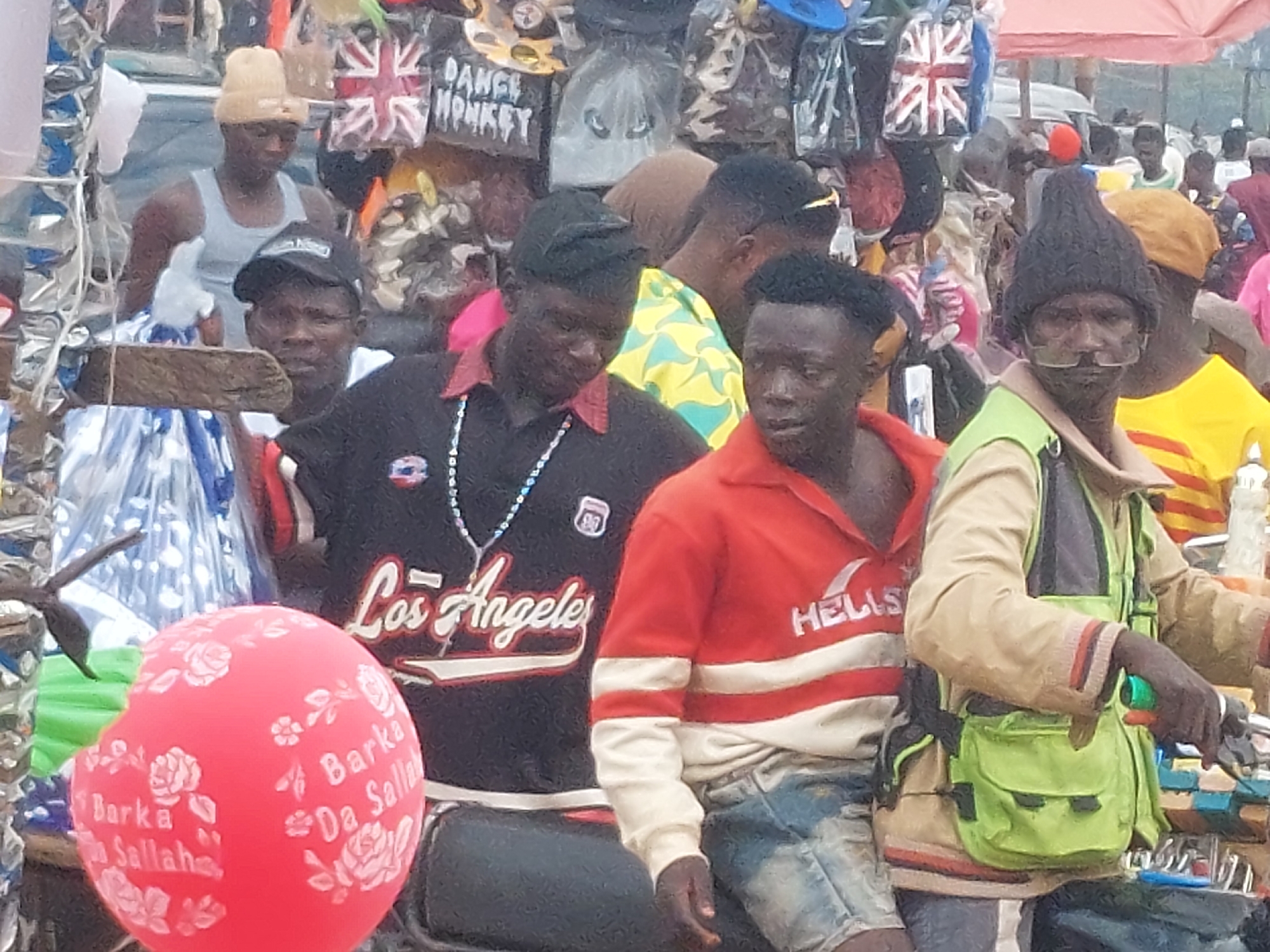 The image depicts an outdoor market scene in Unknown, Unknown, featuring multiple individuals and various merchandise under bright daylight conditions.
In the foreground, three adult males are seated closely on what appears to be a motorcycle or scooter. The individual on the far right, positioned as the driver, wears a dark grey knitted beanie, light-framed eyeglasses or goggles, a beige long-sleeved shirt, and a light green multi-pocketed vest. His right hand grips a vehicle handlebar with a green element. He looks forward and slightly to his right. The middle individual, seated behind the driver as a passenger, wears an orange long-sleeved shirt with two white horizontal stripes on the chest and sleeves, and "HELLS" written in white on the left chest. He wears faded blue denim shorts and looks to his left. The leftmost individual of this group, also a passenger, wears a black long-sleeved shirt with "Los Angeles" in white cursive text and a white "88" emblem on the left chest. He has a long beaded necklace with a cross pendant. He looks downward and to his right. A red balloon with white floral patterns and the text "Barka Da Sallah" is partially visible in the bottom left corner, in front of this group.
Behind the foreground subjects, a bustling market environment is visible. To the left, a light-skinned male wearing a light yellow beanie and a grey tank top stands, looking forward. His left arm is bent upwards. Behind him and to his right, a dark-skinned male wearing a black baseball cap is partially visible, looking forward. Overhead and in the mid-ground, various items of merchandise are displayed, including a bag or pillow featuring the Union Jack flag, another item with the text "DANCE MONKEY," and several plastic-wrapped bags, some with animal face designs. A rough wooden beam horizontally crosses the mid-left portion of the image, possibly part of a stall structure.
In the right background, a man wearing a bright yellow shirt with red horizontal stripes is partially visible, looking to his left. Further back, an orange market umbrella is open. Numerous other indistinct figures and market stalls populate the background, contributing to a crowded atmosphere. The ground is largely obscured but appears uneven in visible sections.