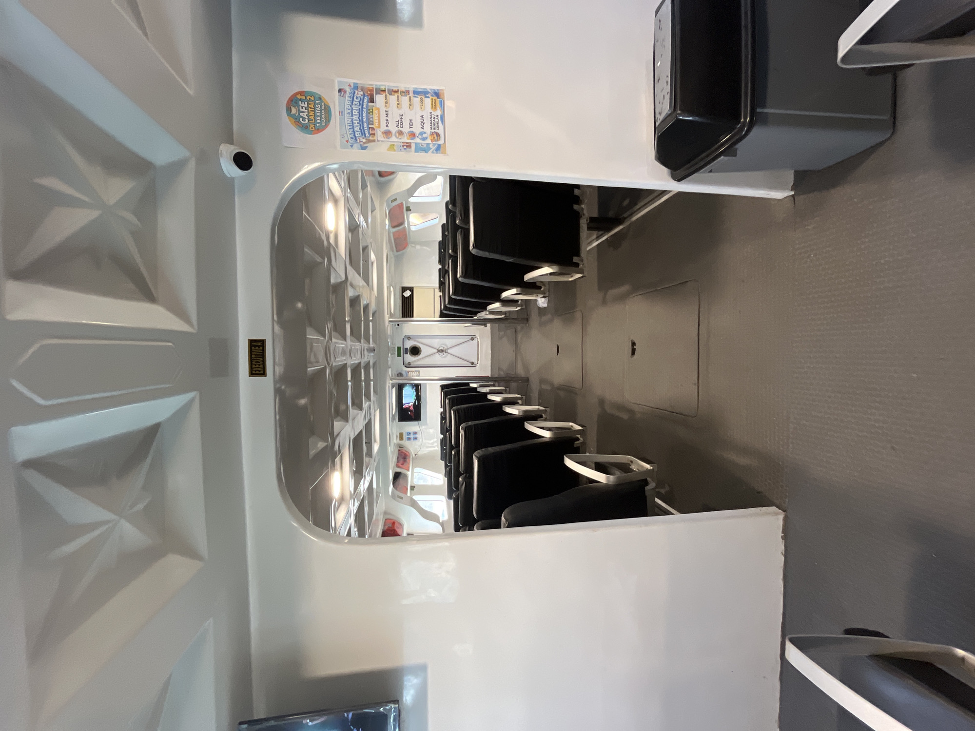 The image shows the interior of a passenger ferry in Karimunjawa, Indonesia. The view is down a central aisle with rows of black, upholstered seats on either side. The seats appear to be arranged in pairs. The ceiling of the ferry is white and features a geometric, faceted design. A security camera is visible on the ceiling near a sign that reads "EXECUTIVE A". To the right, a menu is posted on the wall, advertising items like "POP MIE", "ALL COFFEE", "TEH", "AQUA", and "MAKANAN/MINUMAN TAMBAHAN". The flooring is a dark, textured material. At the end of the aisle, there is a white doorway with a dark border and a window. A black trash receptacle is positioned on the right side of the aisle near the foreground.