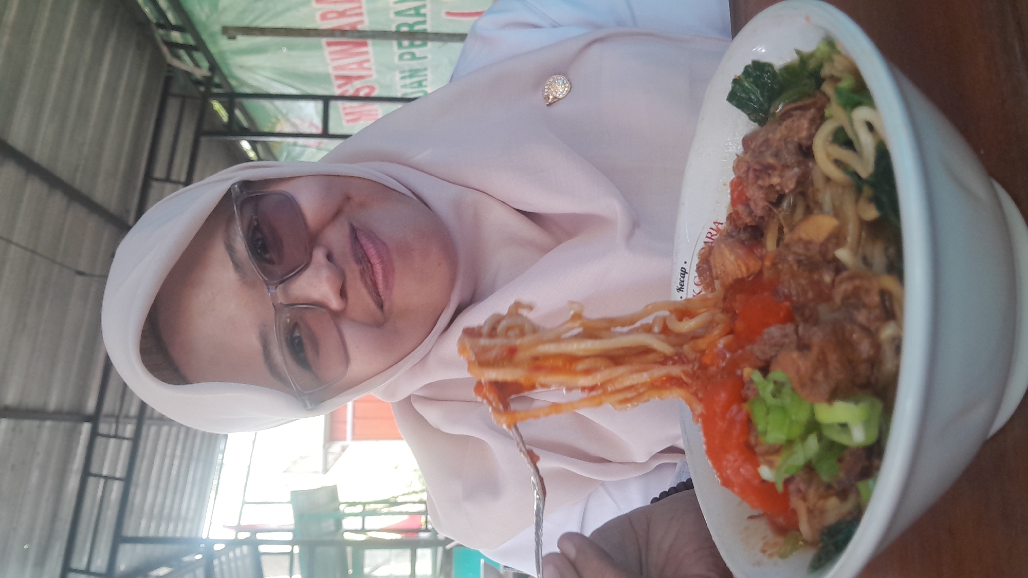 A woman wearing a light pink hijab and glasses is captured in a close-up shot, holding a forkful of noodles. She is smiling and looking directly at the camera. The noodles, coated in a red sauce, are being lifted from a white bowl. The bowl contains more noodles, chunks of meat, and chopped green vegetables. In the background, there are glimpses of an outdoor setting with a metal roof structure and a green banner with Indonesian text. The location is identified as Ngloram, Indonesia.