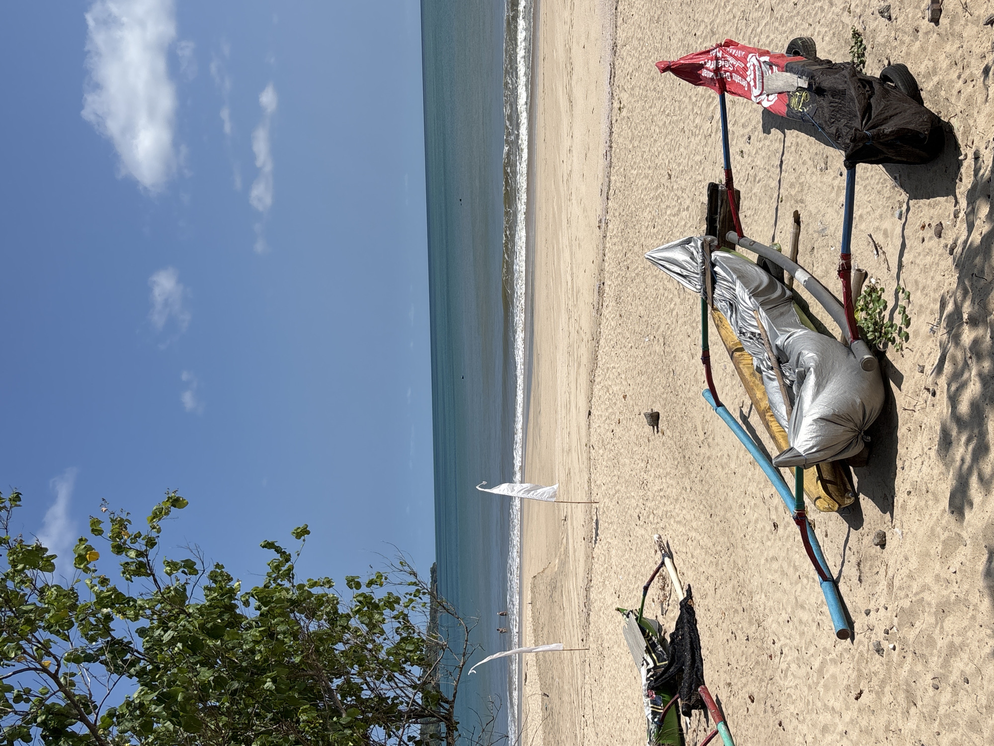 A beach scene in Kuta, Indonesia, shows a shoreline with gentle waves lapping the sand. The sky is blue with scattered clouds. A tree with green leaves stands on the left. On the beach, there's an assortment of objects, including a makeshift shelter with a red-and-white canopy, metallic-colored bags, and various supporting poles. Two small white flags are stuck in the sand near the water's edge. The scene is undisturbed, with no people directly visible.