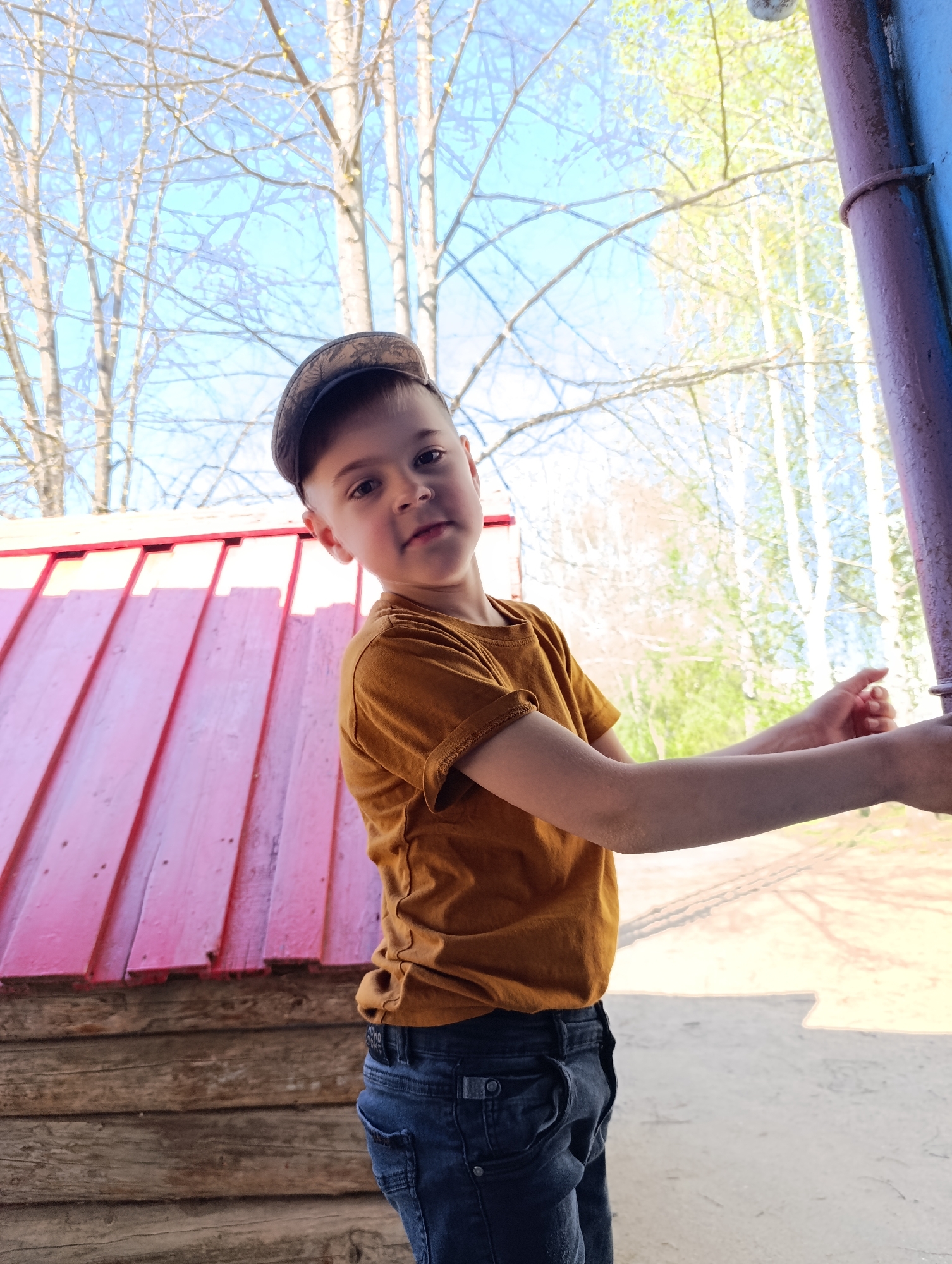 A young boy in a brown t-shirt, jeans, and a cap stands outdoors, likely at a playground. He is holding onto a purple metal pole. To his left is a small wooden structure with a pink roof. The background features trees with sparse foliage against a light blue sky. The location is in Chaikovsky, Russia.