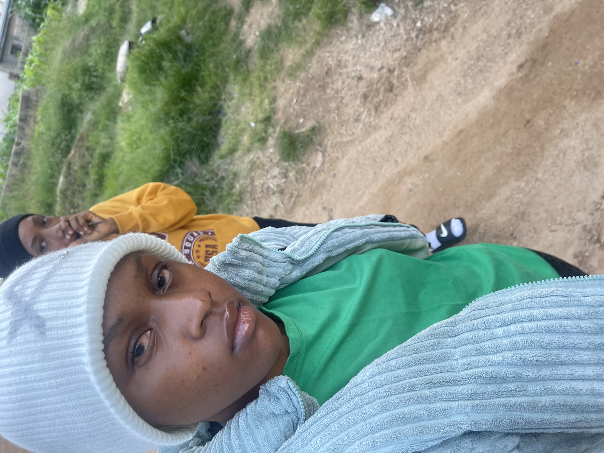 The image shows two young women in an outdoor setting. The woman in the foreground is wearing a light blue corduroy jacket over a green t-shirt and a white beanie. She is looking towards the camera with a neutral expression. Her lips are glossy. Behind her, another young woman is visible, wearing a yellow hooded sweatshirt and a black beanie. She is partially obscured and appears to be holding something to her face. The background consists of a grassy slope with some vegetation and a building structure. The ground in the foreground is a dirt path. The scene is consistent with a casual outdoor environment in a Nigerian city like Rafin Par, characterized by its natural and built-up elements.
