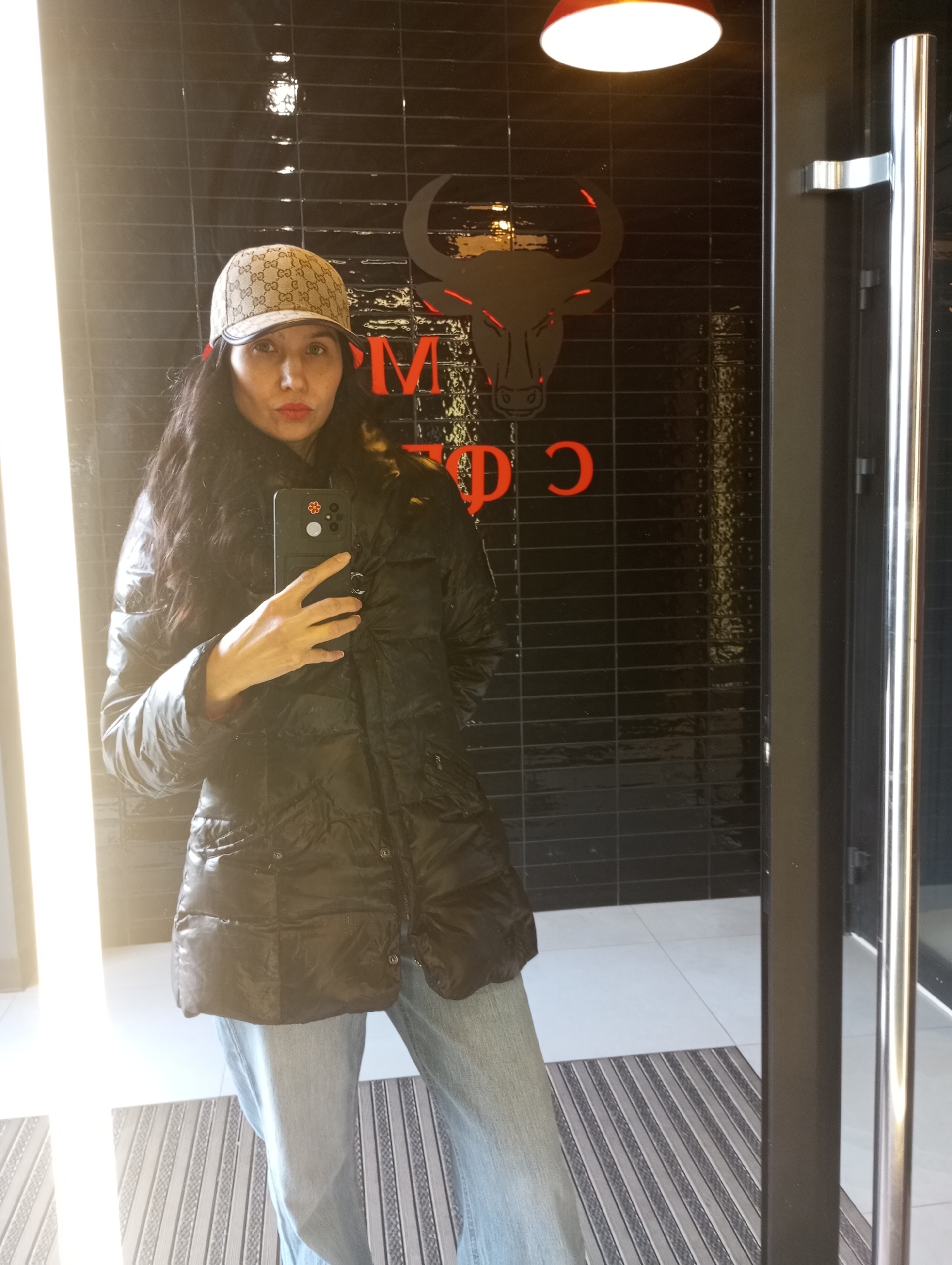 A woman is captured in a mirror selfie, standing indoors in what appears to be an entrance or hallway in Izhevsk, Russia. She has long, dark, wavy hair and wears a beige baseball cap adorned with a prominent Gucci monogram pattern, a black glossy puffer jacket, and light blue jeans. She holds a black smartphone with a decorative flower sticker and a circular element on its back, using it to take the photo. Her lips are accented with red lipstick, and she maintains a neutral expression.
The background features a black tiled wall, upon which a large, stylized black bull head logo with red outlines is displayed, likely representing a business such as a restaurant or cafe. Below the bull's head, partial red lettering is visible, including "M", "D", and "Э". An overhead red and white light fixture casts light from the top right. To the right, a chrome, vertical door handle suggests the presence of a glass door. The flooring comprises white tiles, with a striped doormat placed directly beneath her. A bright vertical light source or reflection illuminates the left side of the image.