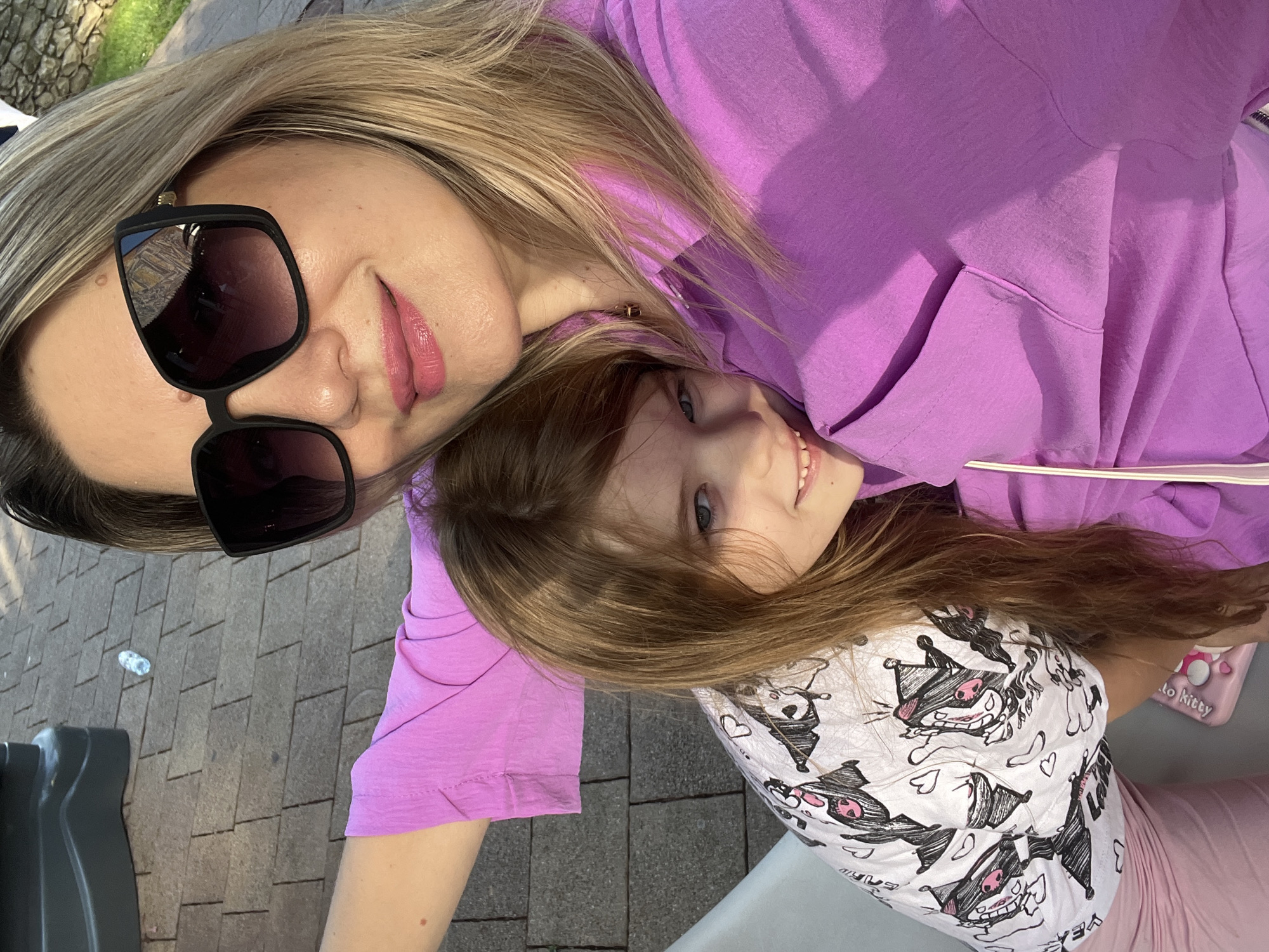 A woman and a young girl are lying down for a selfie outdoors. The woman is wearing a purple hooded sweatshirt and large black sunglasses. Her lips are painted pink, and her blonde hair is flowing. The girl, positioned below the woman, has long brown hair and is looking towards the camera with a slight smile. She is wearing a white t-shirt with a cartoon character print and pink leggings. The background shows a paved surface with rectangular gray tiles, suggesting an outdoor patio or walkway. Sunlight casts shadows, indicating it is daytime, possibly late afternoon or early evening. The overall mood appears relaxed and happy, as they pose together. The scene is outdoors, with hints of greenery and possibly a stone wall in the upper left corner.