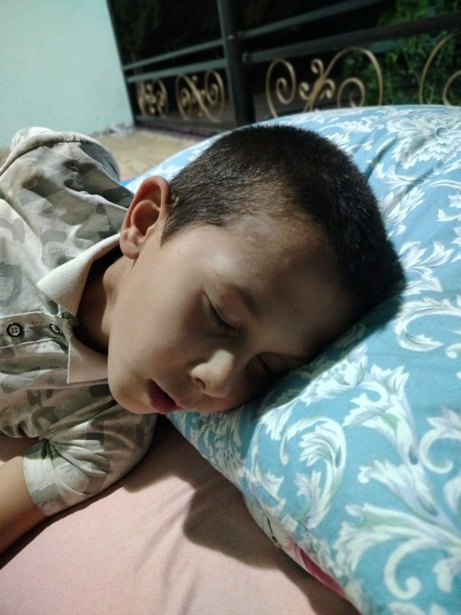 A young boy is sleeping on his side, resting his head on a blue and white patterned pillow. He is wearing a light-colored shirt with a camouflage pattern. The background shows a blurred view of what appears to be an outdoor setting, possibly a balcony or patio, with a railing and greenery. The overall scene suggests a peaceful and restful moment. The image does not provide any specific visual cues to pinpoint its location within the city of Көлкент, Kazakhstan.