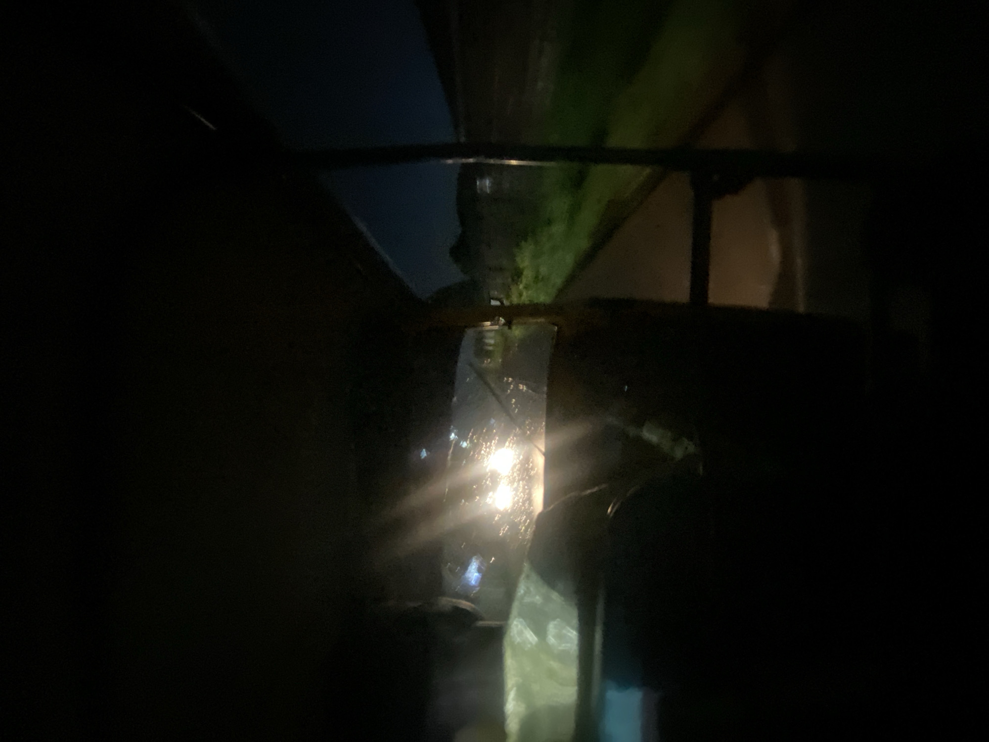 The image is a dark, interior view from the back seat of a vehicle, possibly an auto-rickshaw or tuk-tuk, at night. The windshield is visible with raindrops, and bright headlights from an oncoming vehicle obscure much of the forward view. The interior of the vehicle is mostly black, with faint outlines of the seats and a steering wheel visible. Outside the vehicle, a dimly lit street or road can be seen with green vegetation and some buildings in the distance. The scene suggests movement, likely on a road in a rural or semi-urban area at night, possibly Gura Top, Nigeria, based on the provided context.
