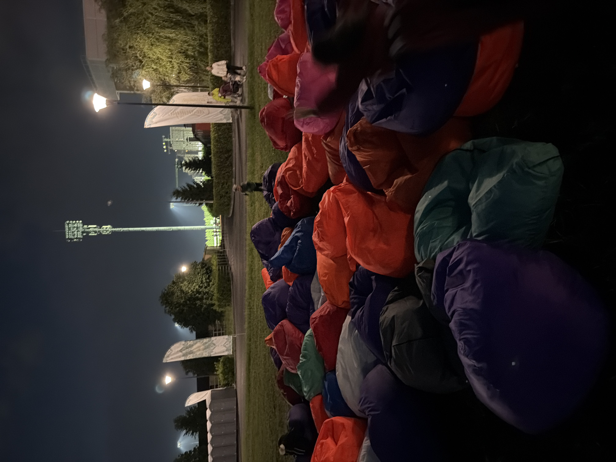 An outdoor scene at night shows a large gathering of colorful beanbag chairs spread across a grassy area. In the background, a tall tower with lights stands against a dark sky, possibly a stadium or communication tower. Several streetlights illuminate parts of the scene, casting a warm glow on the surrounding trees and buildings. A few portable toilets are visible to the left. In the mid-ground, two women are pushing a stroller, while a young child walks in front of them. The beanbag chairs, in various shades of red, orange, purple, and green, are piled up and ready for use, suggesting an event such as an outdoor movie screening or a festival. The overall atmosphere is relaxed and inviting.