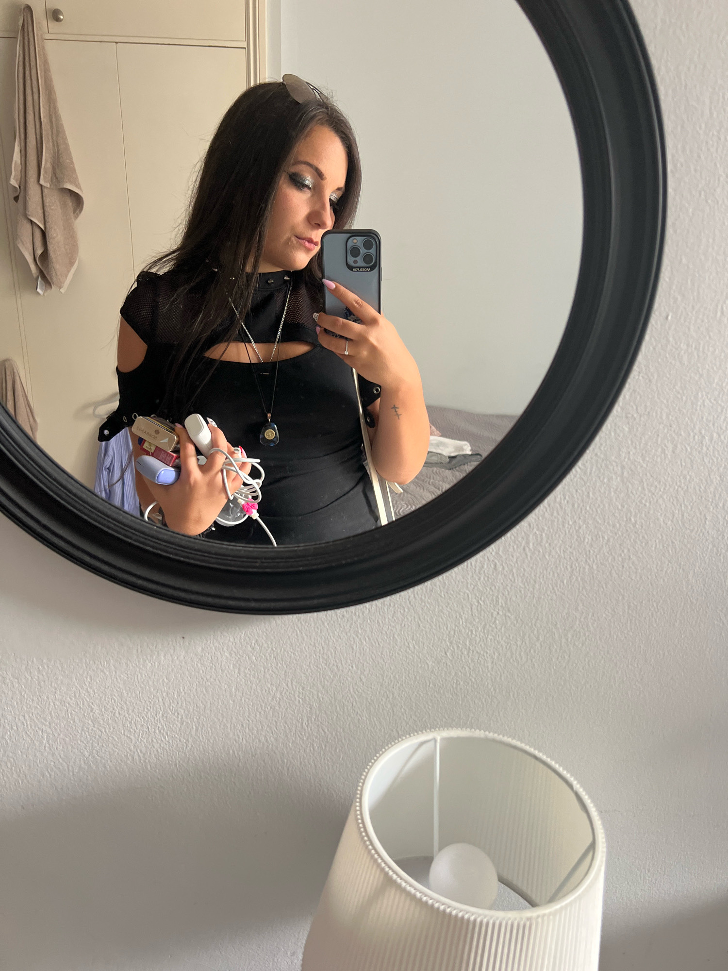 A woman with long, dark hair and dramatic eye makeup is taking a mirror selfie in a room. She is wearing a black, cut-out top and is holding a collection of items in her left hand, including what appear to be electronics and cables. A silver necklace with a dark pendant is visible. Reflected in the mirror, a towel hangs on a wardrobe to the left, and a bed is visible in the background to the right. The room appears to be a bedroom or dressing area. A white lampshade with a lightbulb is in the foreground. The image is taken in a private residence in London, United Kingdom.