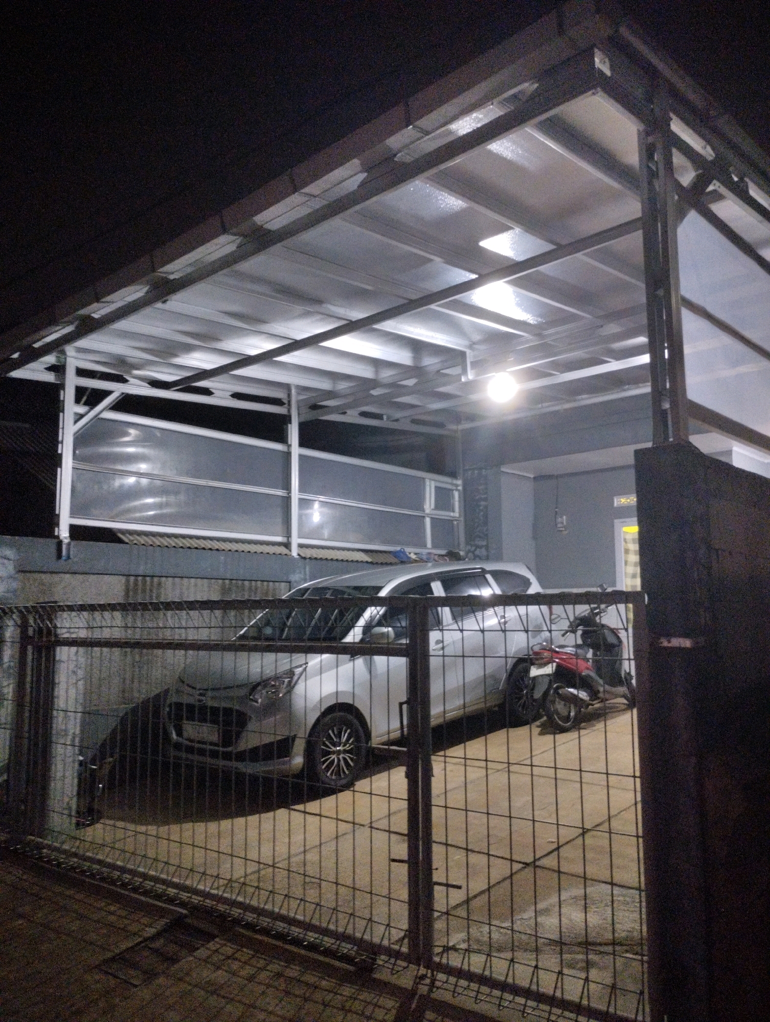 In Cimenyan, Indonesia, a silver hatchback car and two motorcycles are parked under a translucent, corrugated metal canopy. The canopy is supported by a metal frame and is illuminated by overhead lights. A wire fence runs across the foreground, partially obscuring the vehicles. The ground beneath the vehicles is paved with concrete tiles. The background shows a gray wall with a yellow-tinted window and a dark, textured surface on the right. The overall scene appears to be a residential or private parking area at night.
