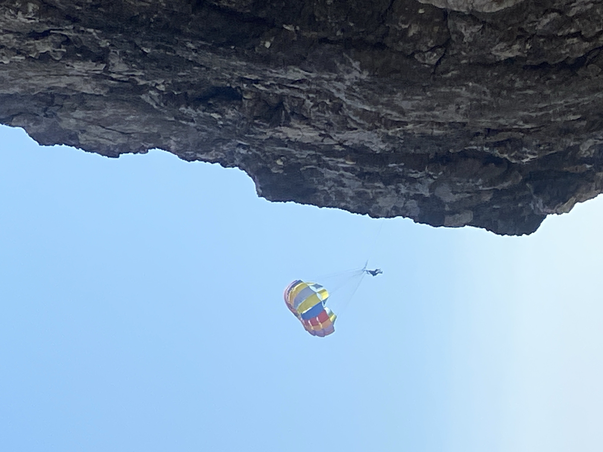 A parasailor is in mid-air against a clear blue sky, attached to a colorful parachute. The parasailor appears as a small, dark silhouette suspended from the parachute by thin lines. The parachute is a mix of red, yellow, blue, and white panels, billowing in the wind. To the right of the parasailor and occupying the majority of the frame is a massive, rugged, dark gray rock face. The rock is textured with numerous cracks, fissures, and irregularities, suggesting a natural cliff formation. The scene is set outdoors, likely near a coastal area given the presence of the cliff. The overall impression is one of adventure and outdoor recreation against a dramatic natural backdrop.