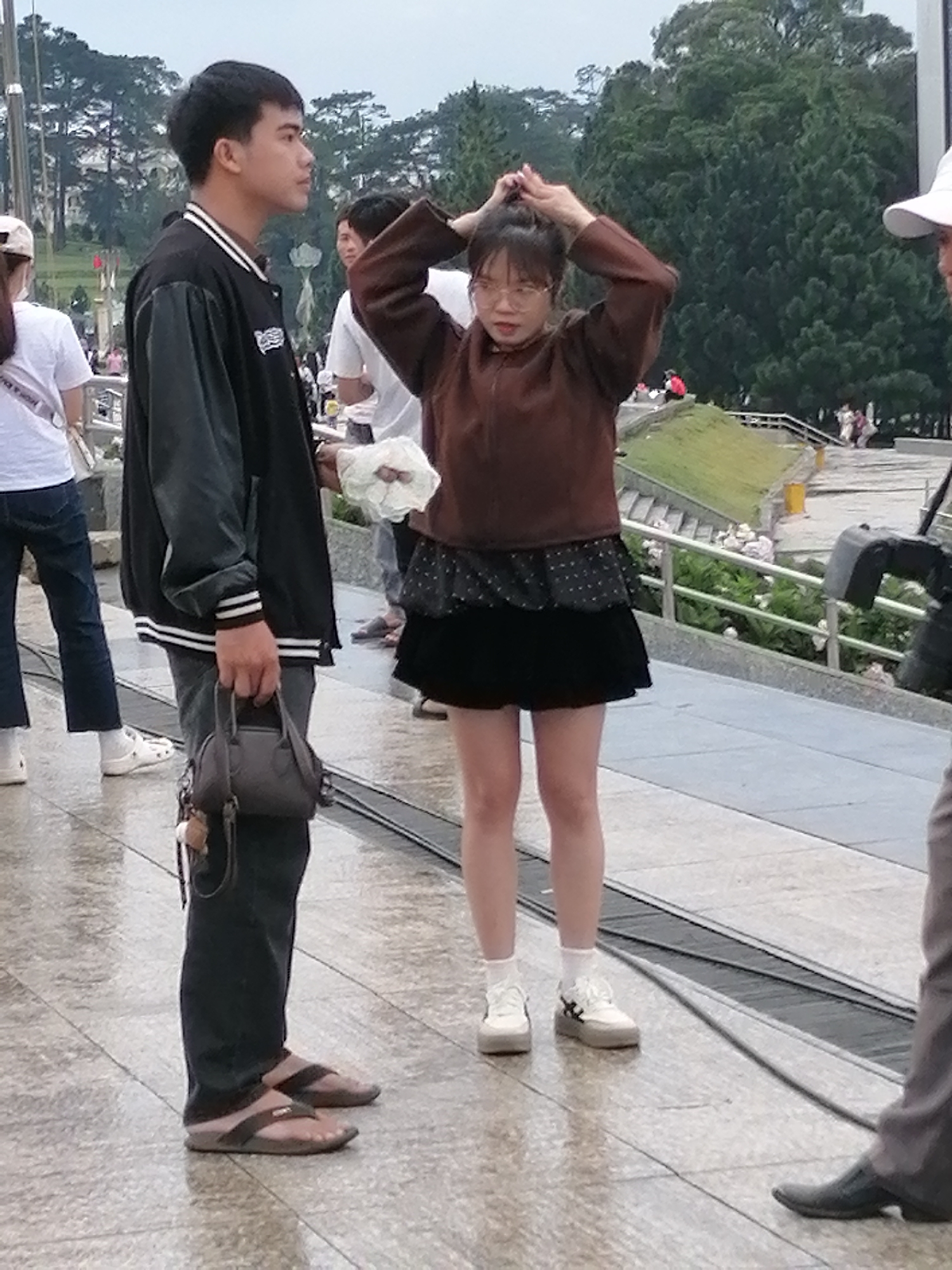 The image depicts an outdoor scene in Da Lat, Vietnam. In the foreground, a young Vietnamese man stands with his side facing the camera, holding a dark grey handbag in his right hand. He is wearing a black bomber jacket with white striped cuffs, dark pants, and brown flip-flops. To his right, a young Vietnamese woman is facing the camera, her hands raised to her hair. She is wearing a brown long-sleeved shirt, a black skirt with a white polka dot pattern, white socks, and white sneakers. The ground appears wet, suggesting recent rain. In the background, a lush green hillside with trees and stairs can be seen, along with other individuals and a railing. A portion of a camera rig is visible on the right side of the frame. The overall atmosphere suggests a public space in Da Lat.
