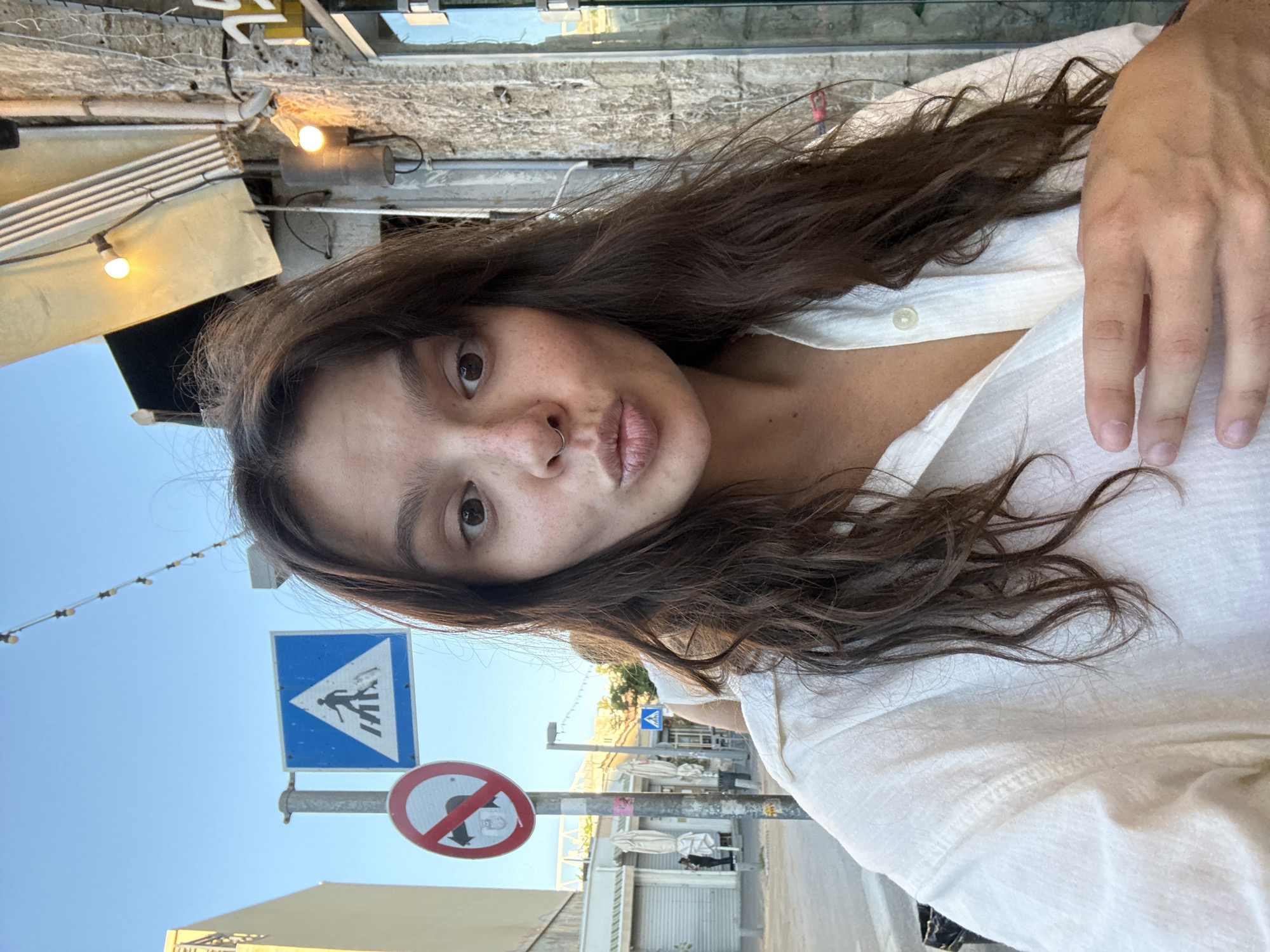 In Tel Aviv, Israel, a woman with brown hair and freckles is taking a selfie outdoors. She is wearing a white button-up shirt. She has her lips puckered as if to kiss and her left hand is on her chest. Behind her, a blue pedestrian crossing sign and a red circular sign with a diagonal line through it are visible. The background shows a street scene with buildings and some people. The lighting suggests it is daytime.