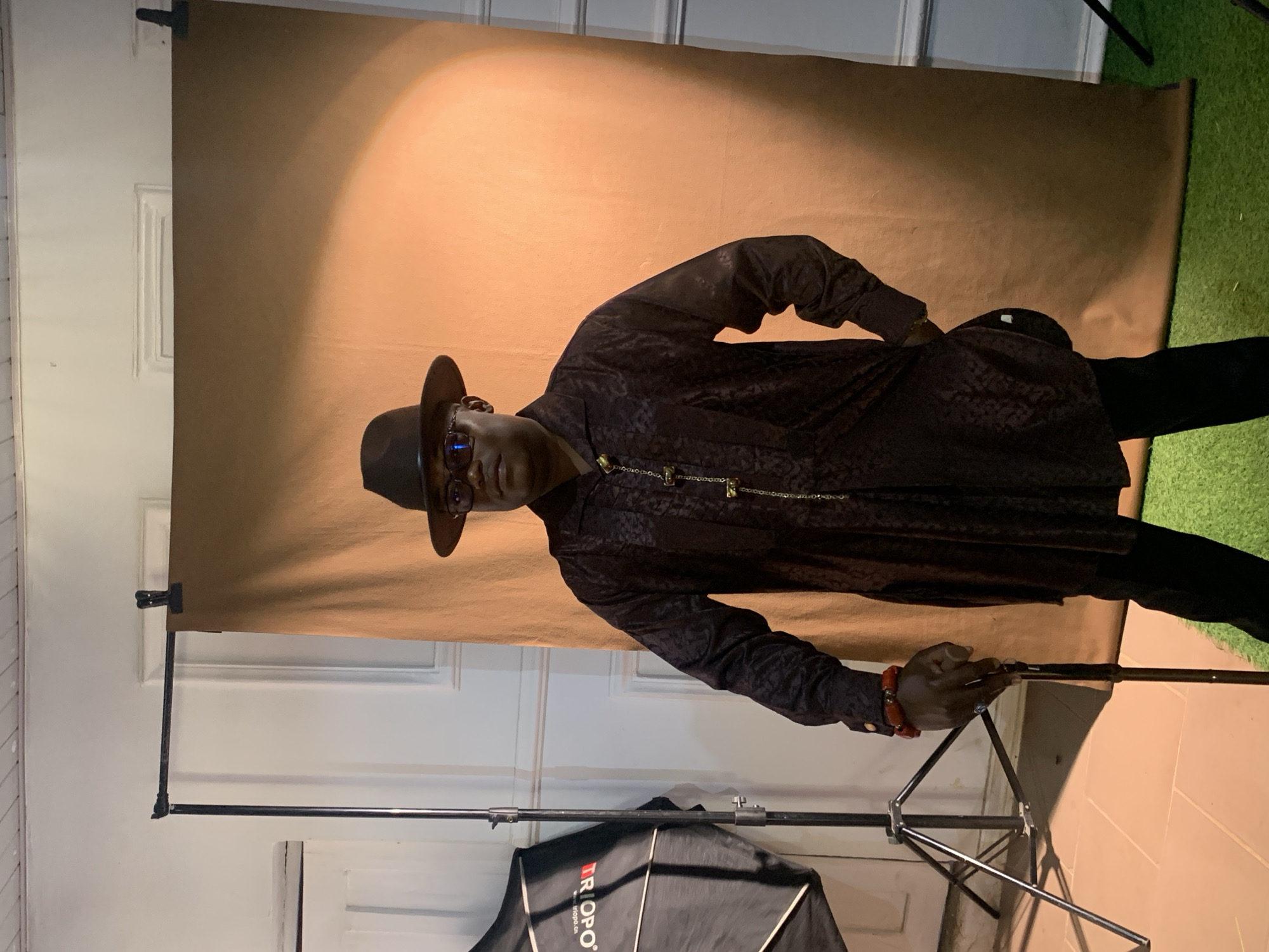A person is posing indoors in front of a brown backdrop with artificial green grass in the upper right corner. The person is wearing a dark patterned long-sleeved shirt, black pants, a hat, and glasses. They are holding onto a black tripod stand. A softbox is visible in the lower portion of the image. The location is likely a studio or indoor space in Jos, Nigeria.