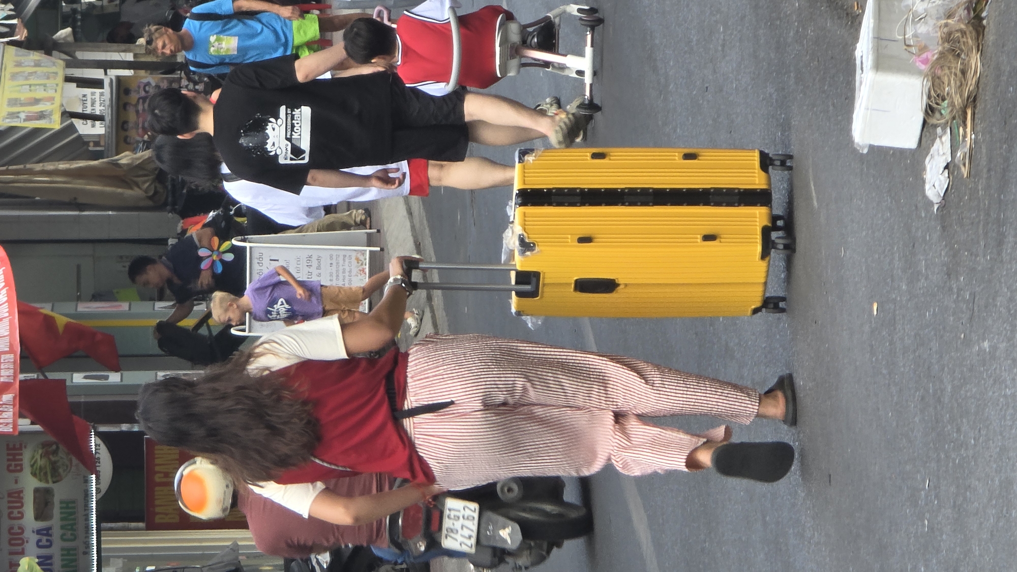 A bright daylight street scene captures a moment of daily life in an urban area of Vietnam. In the immediate foreground, a person with long dark hair, seen from behind, is walking away from the camera while pulling a distinctive, large bright yellow hard-shell suitcase with its handle extended. This individual is dressed in a red short-sleeved top and light-colored, vertically striped trousers, paired with dark sandals. To their left, a parked motorcycle or scooter is visible, bearing the Vietnamese license plate "78-G1 247.62".
Behind the person with the suitcase, another individual walks, clad in a black t-shirt featuring a white "Kodak" logo and black shorts. Further into the midground, several other people are visible, including a person wearing a black t-shirt with a colorful flower design, a child in a purple t-shirt, and a person in a blue t-shirt and green shorts. Some debris and what appears to be discarded packaging lie on the pavement to the right.
The background is lined with various storefronts or stalls adorned with multiple signs displaying Vietnamese text. Notable text includes "LỌC CỦA - GHẸ" (likely referring to crab or seafood dishes), "BÁNH CANH" (a type of Vietnamese noodle soup), and service advertisements for "CỘI ĐẦU" (hair wash/shampoo) indicating prices "từ 49k" and operating hours "8:30 - 18:00". Red flags with yellow stars, characteristic of the Vietnamese national flag, are also displayed, reinforcing the location context of Nha Trang. The scene depicts a lively, everyday street environment, possibly featuring tourists or travelers among the local populace.