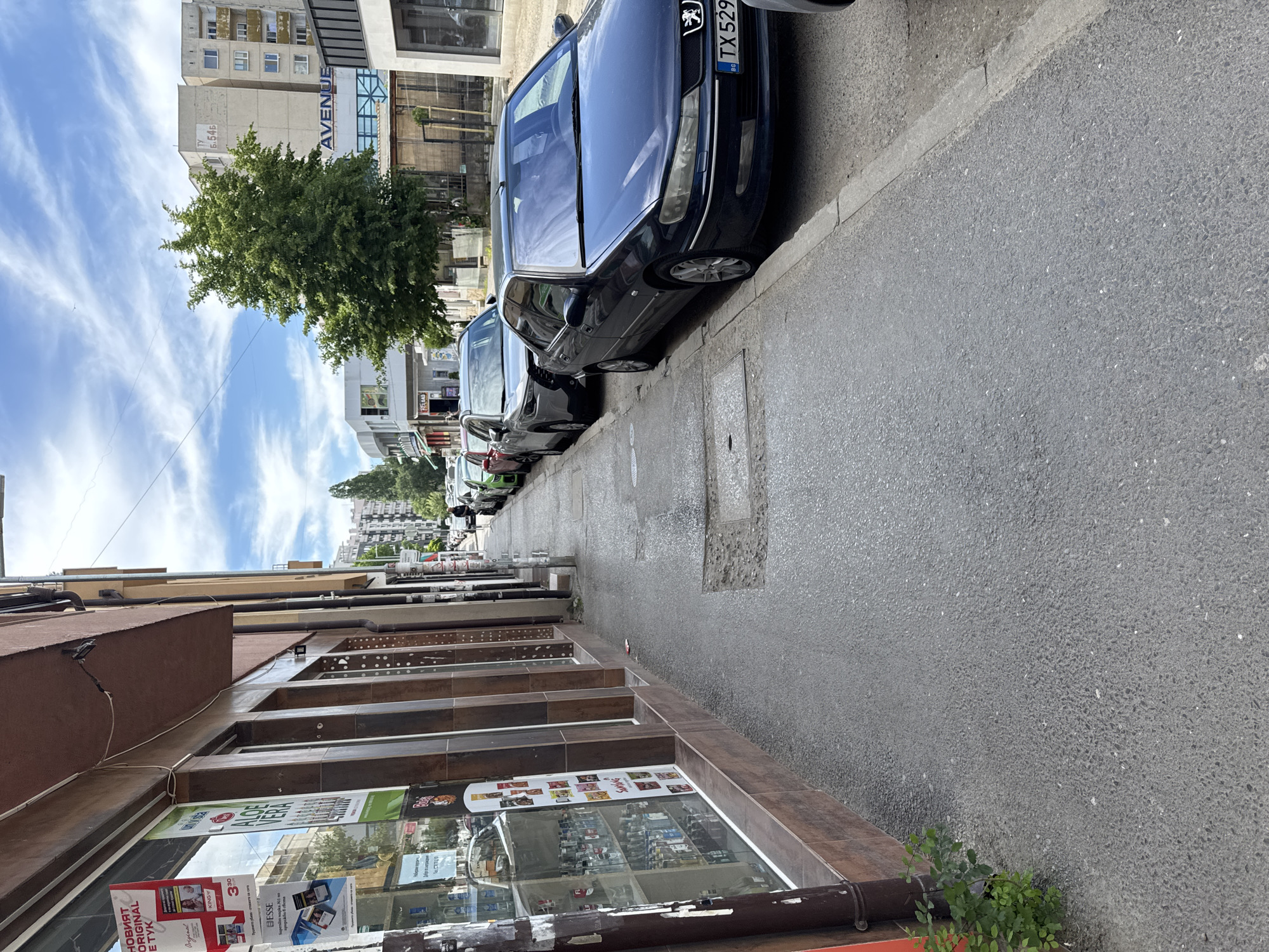 The image depicts a street scene in what appears to be Sofia, Bulgaria. A row of parked cars lines the right side of the frame, with a dark blue Peugeot visible in the foreground, license plate "TX 529".
The street appears to be a mix of residential and commercial, with buildings visible in the background and a shopfront along the left side of the frame. Several tobacco-related advertisements are visible in the shop window. The building in the background has the text "AVENUE" written on it, and the text "TY Б.54Б" (likely an apartment number or address) is visible near the top of the building.
The sky is partially cloudy, with a bright blue sky visible behind the buildings and trees. The pavement appears to be wet.
