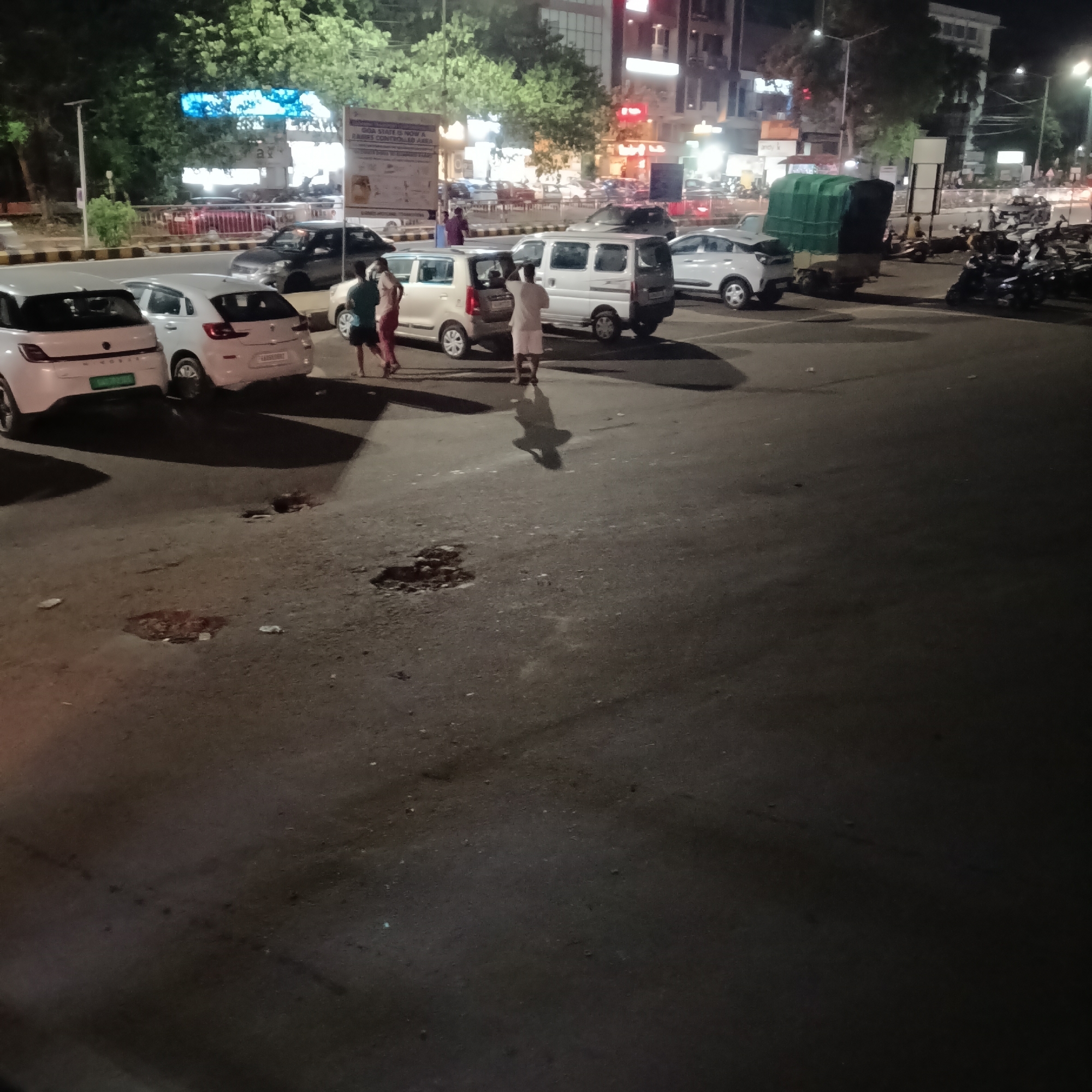 The scene is a nighttime view of a parking area in Margao, India. Several cars and motorcycles are parked along a road with a center median. Two people are walking between parked cars, while another person is walking away from the vehicles. Streetlights illuminate the area, and buildings are visible in the background, including at least one establishment with a red illuminated sign. The road surface in the foreground has visible potholes.