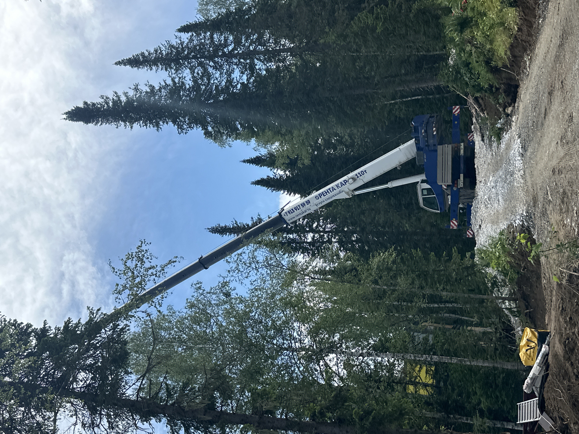 A large blue and white crane is positioned on a dirt road in a dense forest. The crane's boom extends upwards, reaching towards the top of a tall evergreen tree. The tree appears to be leaning or in the process of being lowered. The area around the crane is uneven, with piles of gravel and dirt. Various equipment, including what appears to be lumber and a yellow bag, are scattered on the ground near the base of the slope. The background is filled with a canopy of tall evergreen trees. The sky is partly cloudy. The scene is indicative of tree removal or logging operations. The location is identified as Sheregesh, Russia.