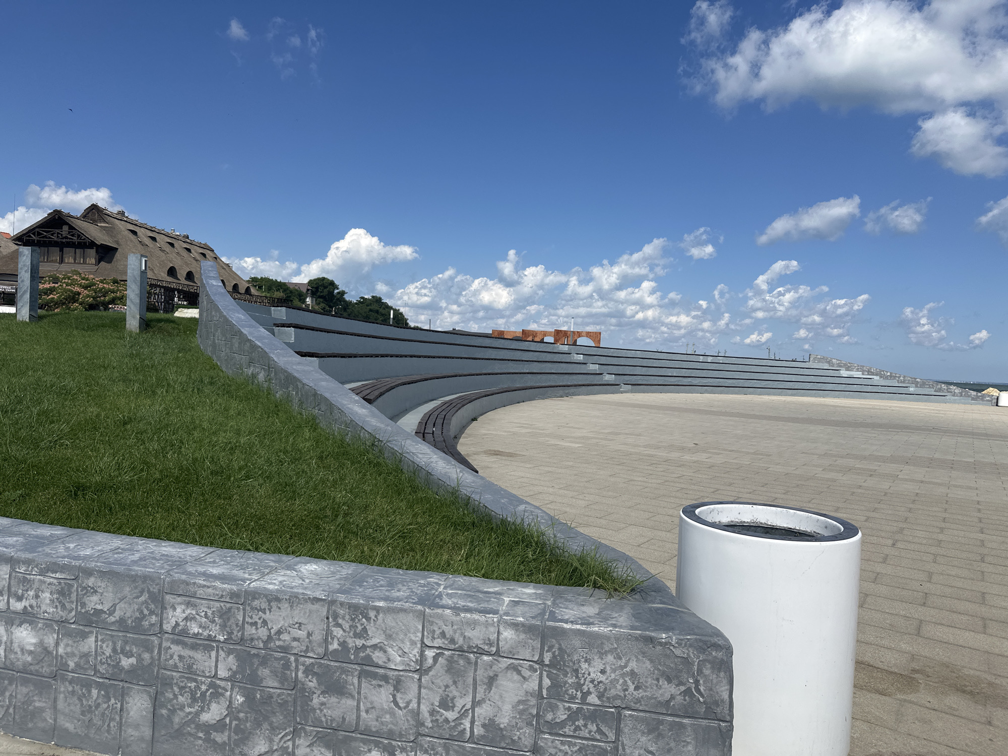 The image depicts an outdoor amphitheater with tiered seating, located in the city of Taman, Russia. The amphitheater is built along a curved embankment, with the seating area constructed from dark wooden planks and grey concrete borders. A patch of vibrant green grass slopes upwards to the left of the seating area, bordered by a textured grey stone wall.
In the background, a large building with a thatched roof, characteristic of traditional Russian architecture, stands prominently. The building appears to be a restaurant or a similar establishment, with visible windows and wooden beams. Beyond the building, a line of trees can be seen, suggesting a vegetated area.
The foreground of the image features a paved walkway made of light-colored rectangular tiles. A white cylindrical trash can with a black rim is placed on the walkway, to the right of the stone wall. The sky above is a clear, bright blue, dotted with fluffy white clouds. The overall scene conveys a sense of public space designed for gatherings and performances, set against a backdrop of natural and architectural elements.