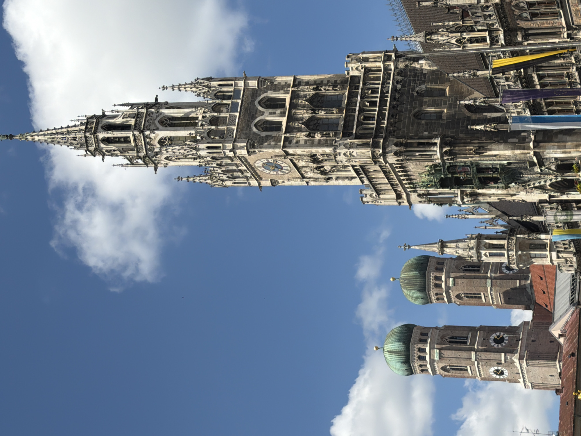The image captures a view of the New Town Hall (Neues Rathaus) and the Frauenkirche in Munich, Germany, under a bright blue sky with scattered clouds. The New Town Hall's prominent Gothic tower, featuring intricate stonework, gargoyles, and a large clock face, dominates the right side of the frame. To the left, two towers of the Frauenkirche, characterized by their distinctive green onion domes and rose-colored brick, are visible. The foreground includes parts of red-tiled roofs and banners, one of which displays the colors blue, yellow, and white, and another with blue and white stripes. The overall scene is a detailed architectural study of these historic landmarks.