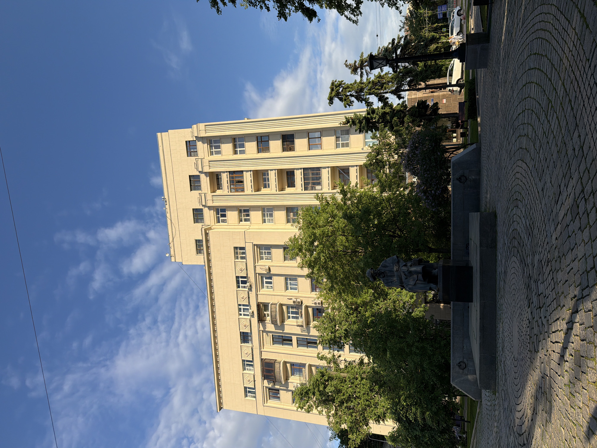The image captures a multi-story building with a symmetrical facade, constructed in a pale, possibly Art Deco-influenced style. The building features evenly spaced windows, some with small balconies. The sky is blue with scattered clouds. Trees partially obscure the base of the building and what appears to be a statue on a pedestal. The ground is paved with cobblestones. This scene is located in Moscow, Russia.