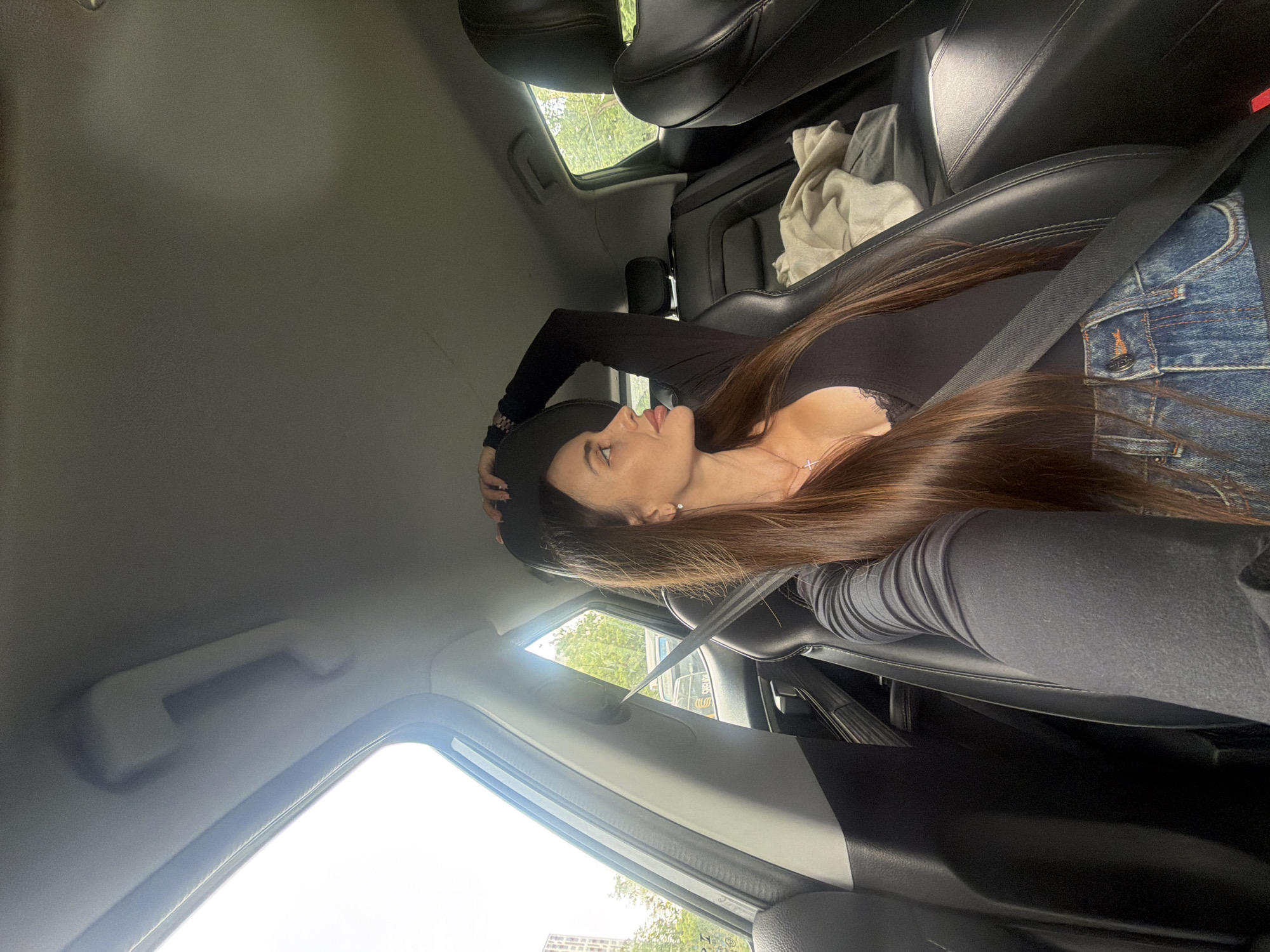 A woman with long, dark brown hair is seated in the passenger seat of a car, looking out the window. She is wearing a black long-sleeved shirt and blue denim jeans. A seatbelt is fastened across her chest. The interior of the car, including black leather seats and part of the dashboard, is visible. Outside the car, trees and buildings are visible, suggesting an urban or suburban environment. The image appears to have been taken during daylight.
