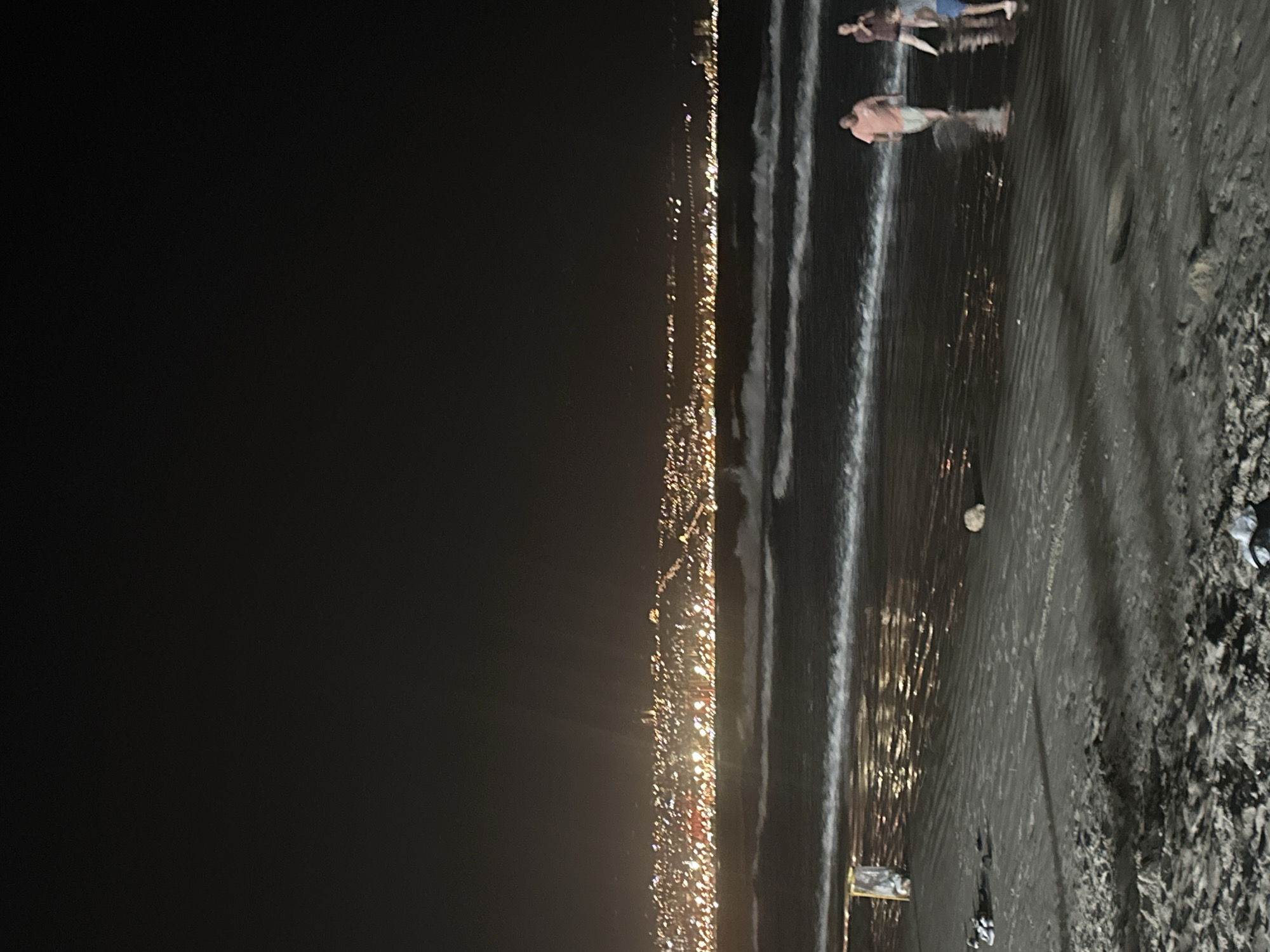 This is a night photograph of a beach, likely in Kiryat Yam, Israel, as indicated by the city lights visible in the background. The sky is dark and clear, with a faint glow from the distant city illuminating the horizon. The sea is calm with gentle waves breaking on the shore, reflecting the city lights. Several people are walking along the wet sand at the edge of the water. One person in a pink shirt and white shorts is walking towards the left side of the frame, with two other figures further to the right, one in a dark shirt and the other in a blue shirt. There are some scattered objects on the sand, including what appear to be shoes or flip-flops on the left side, and a round rock near the center. The sand is dark and appears wet and compacted. The overall impression is of a peaceful evening stroll on the beach.