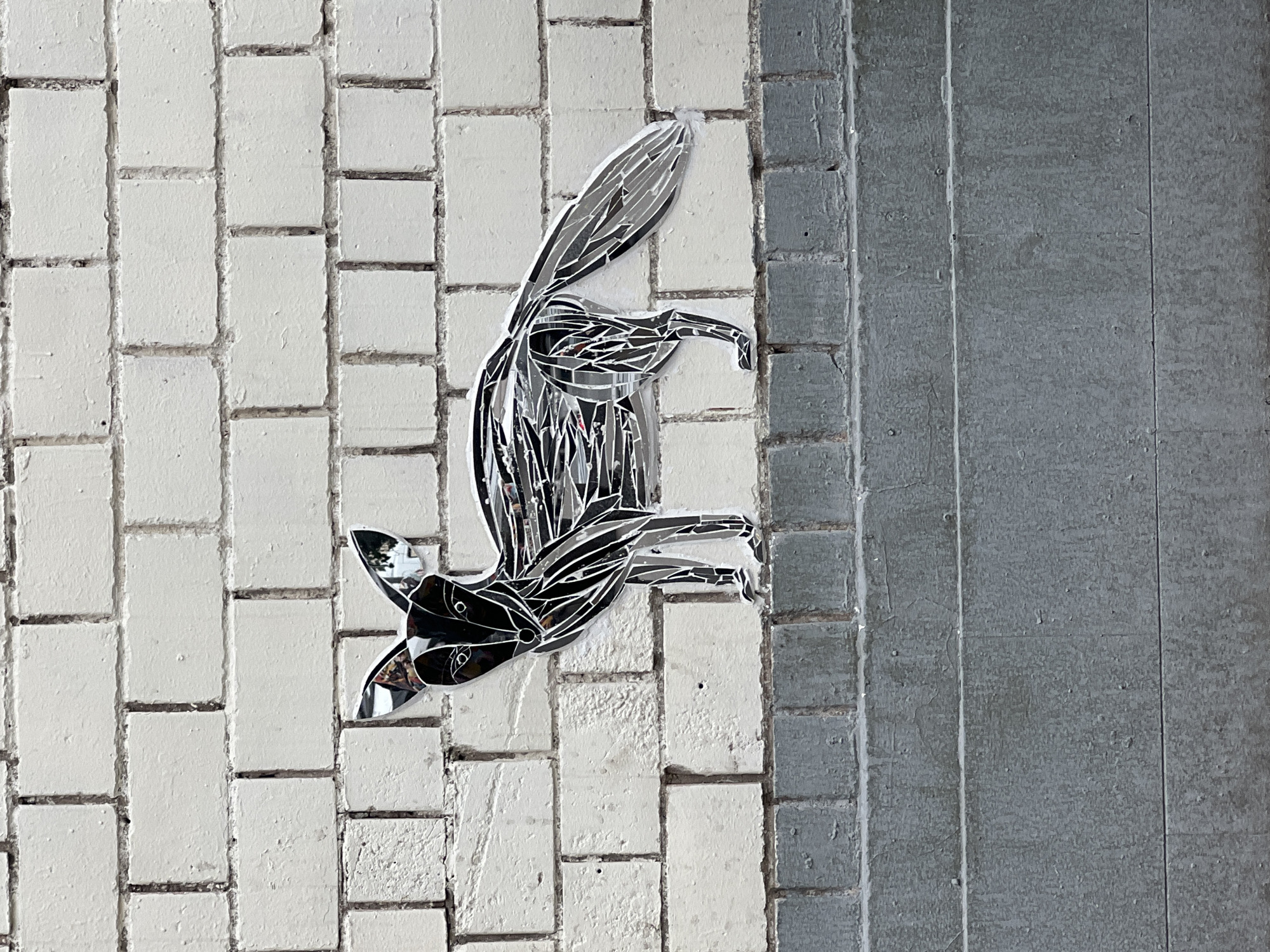 The image shows a mosaic of a fox on a brick wall in Moscow, Russia. The mosaic is made of reflective pieces that create a shimmering effect. The fox is black and white, with large ears and a bushy tail. It is standing on a ledge, and the brick wall behind it is painted white. The lower part of the image shows a grey concrete surface, possibly stairs. The overall scene appears to be an outdoor urban environment.