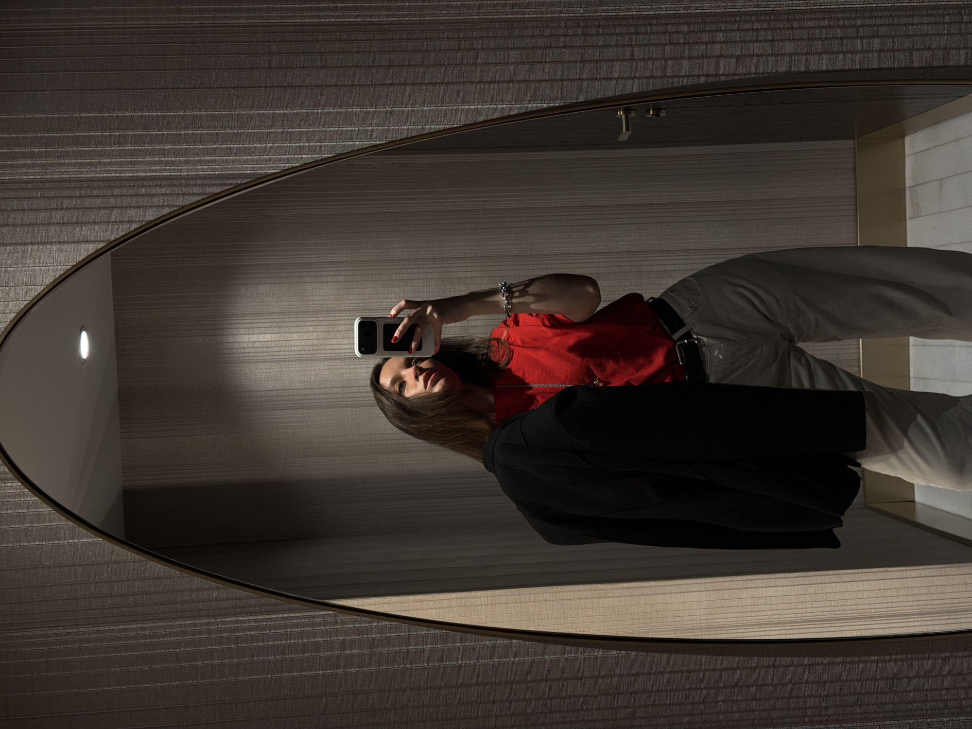 An interior location within Budva, Montenegro. A female subject is captured in reflection, positioned centrally within a large, arch-top mirror with a dark frame. She is taking a selfie, holding a light-colored smartphone with a dark camera module in her raised right hand. Red nail polish is visible on her fingernails. Her attire consists of a red short-sleeved top, light-colored wide-leg trousers, and a dark belt. A black jacket is draped over her left arm. She wears a multi-beaded bracelet on her right wrist and a necklace with a pendant. The surrounding walls, visible directly and in reflection, feature a dark, vertically textured or striped covering. A single recessed ceiling light source is apparent in the reflection. The floor is light-toned, and a door with a brass-colored handle is partially visible to the right of the mirror.