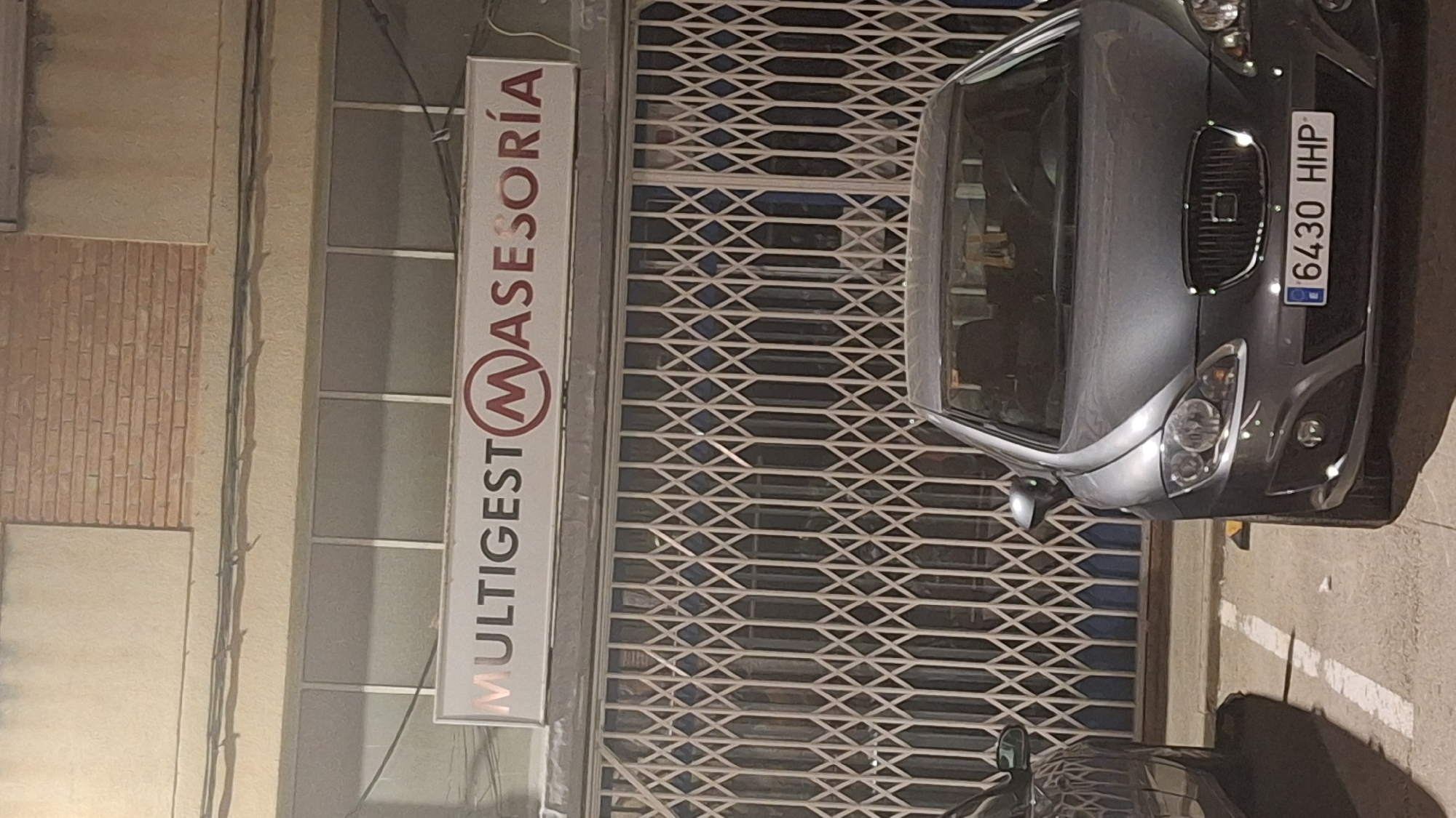 The image shows the exterior of a building with a sign that reads "MULTIGEST MASESORÍA". The building has a rolled-down metal gate in front of a windowed section. A grey car is parked in front of the gate, and part of another car is visible on the left. The license plate of the grey car reads "6430 HHP". The environment appears to be a commercial area or parking lot at night. Based on the context of the sign "MULTIGEST MASESORÍA", this is likely a business establishment in Híjar, Spain.