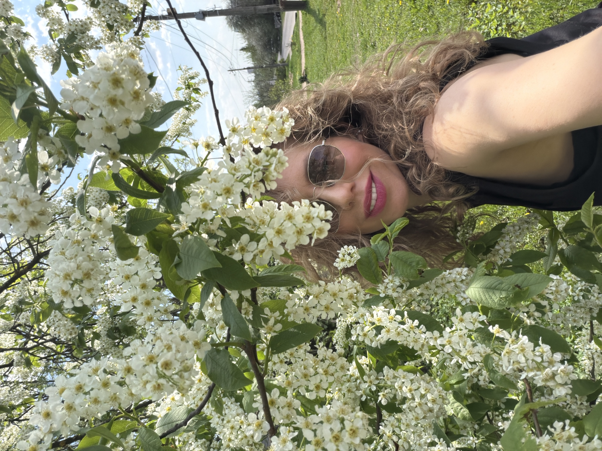 A fair-skinned woman with curly blonde hair and wearing sunglasses is smiling into the camera while lying on the grass near a tree with white blossoms. The location appears to be outdoors, with grass, trees, and utility poles visible in the background. The specific location in the city of Unknown, Unknown, is not discernible from the image.