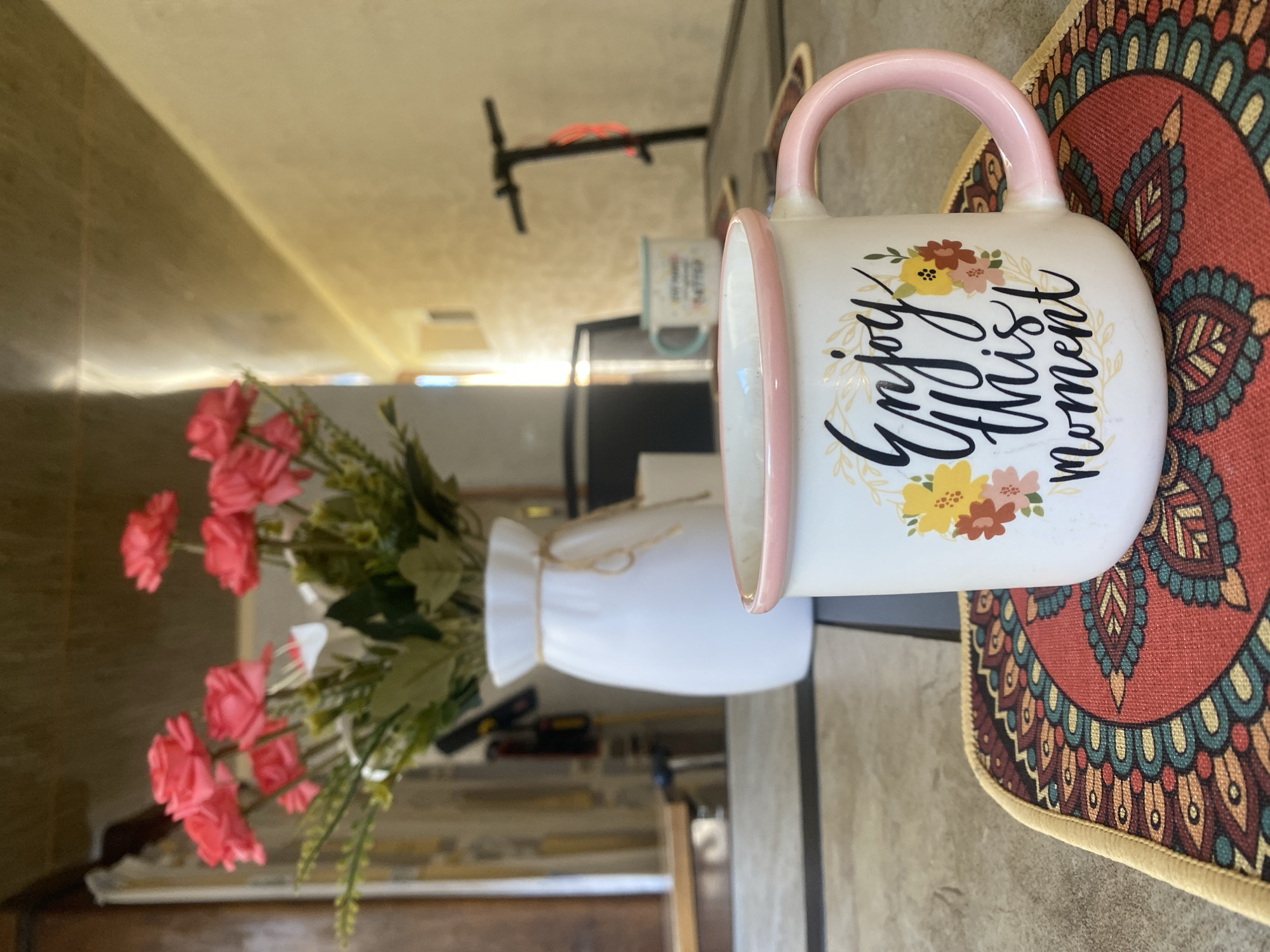 In an indoor setting, a white ceramic mug with a pink rim and handle rests on a decorative placemat. The mug features a floral design and the script "Enjoy this moment." Behind the mug, a white vase holds a bouquet of artificial pink roses and greenery. Further in the background, another mug with a blue rim and a phrase written on it is visible, along with a black scooter. The environment appears to be a room with soft lighting, possibly a dining area or living room. The image does not provide any information suggesting the location is the city of Lagodekhi, Georgia.