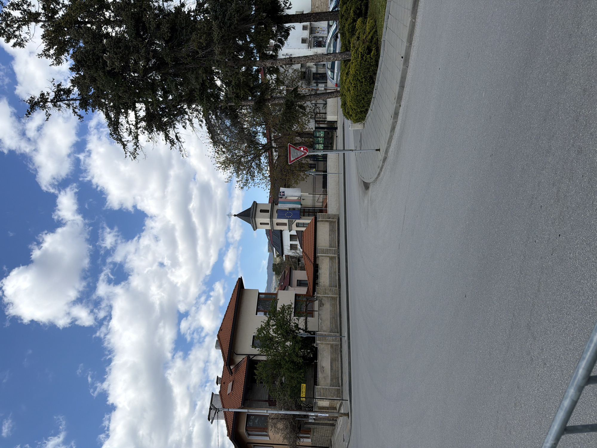 The image depicts a sunlit street scene in Panagyurishte, Bulgaria, under a vibrant blue sky dotted with scattered white cumulus clouds. The perspective is from the roadside, looking down a gently curving asphalt road.
To the left of the road, a sidewalk runs alongside various buildings and natural elements. A prominent building with a distinctive, dark-roofed tower stands out, adorned with multiple flags, including what appears to be the national flag of Bulgaria (white, green, red horizontal stripes) and the flag of the European Union (blue with a circle of yellow stars). Other buildings along the street feature traditional red tile roofs and light-colored stucco or stone facades. Several mature trees, both coniferous and deciduous, provide greenery and shade, their branches reaching towards the sky. Bushes and a black metal fence separate the properties from the sidewalk.
A clear "Give Way" road sign, a red triangle with a white inverted triangle, is prominently displayed on a pole by the roadside. Further down, near the bottom left of the image, a small yellow sign with black Cyrillic text is visible, though the specific words are indistinct. The road itself appears to be empty of vehicles and pedestrians.
The time of day is clearly daytime, likely mid-morning or early afternoon, given the bright sunlight and discernible shadows. The weather is clear and sunny. The architectural style, the specific road sign, and the flags reinforce the given location context of Bulgaria. No specific activity or event is taking place; the scene portrays a quiet, everyday street.