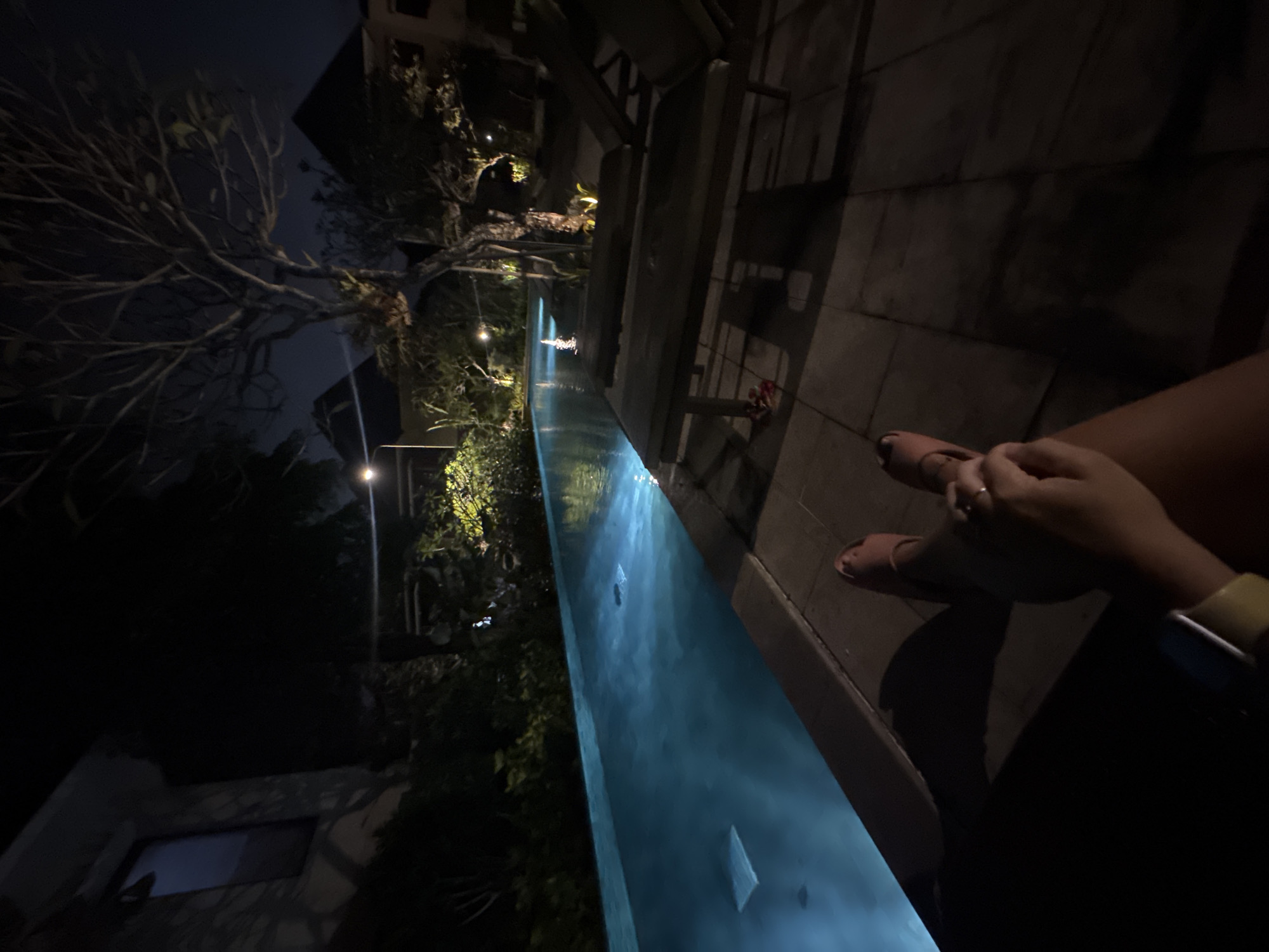 At night in Kuta Selatan, Indonesia, a narrow, blue-lit swimming pool stretches into the darkness. On the right, a person sits on the pool's edge, their feet dangling above the water. They wear pink sandals, and a watch is visible on their wrist. The pool is bordered by stone tiles. Beyond the pool, lush vegetation is illuminated by sporadic lights, and a building is partially visible. A bare tree reaches into the night sky on the left. The scene is dark, with artificial lighting providing the main source of illumination.