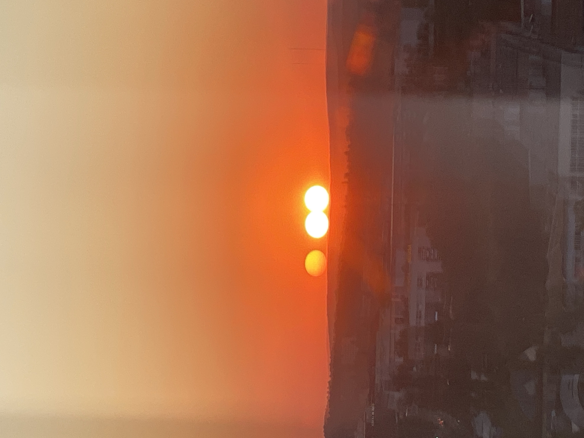 The image captures a striking optical phenomenon: a triple sunset or sunrise observed over the cityscape of Chișinău, Moldova. The sky is dominated by a gradient of warm orange and yellow hues, transitioning from a lighter tone at the top to a deeper orange near the horizon. Against this vibrant sky, three distinct luminous orbs are visible, appearing as multiple suns. The leftmost orb is smaller and appears to be a single sun. To its right, two larger orbs are positioned closely together, possibly appearing as a double sun or a sun being eclipsed by another celestial body. These orbs are positioned above a silhouetted landscape of rolling hills and the indistinct shapes of buildings in the foreground, suggesting an urban or semi-urban setting. The overall impression is one of an unusual atmospheric event, creating a surreal and visually captivating scene.