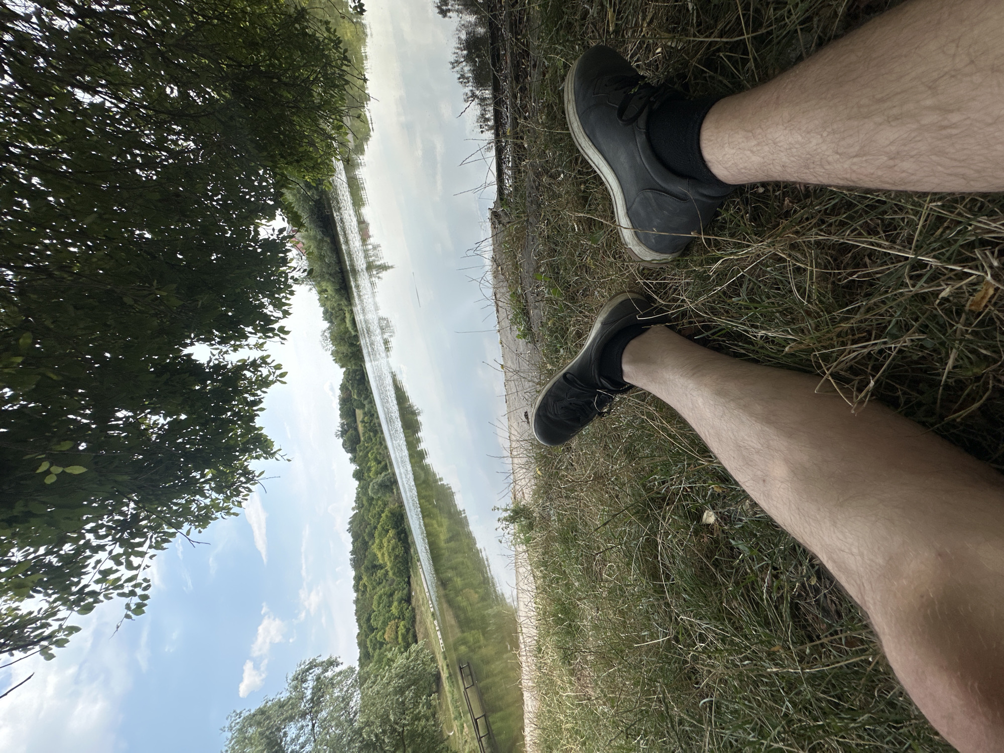 The image depicts a person's legs and feet resting on dry grass by the edge of a calm lake. The person is wearing dark sneakers. The lake is surrounded by lush green trees and vegetation, with a clear blue sky overhead. The reflection of the trees and sky is visible on the water's surface. A small wooden pier or walkway extends into the lake from the left side of the frame. The scene appears to be a natural outdoor setting, possibly a park or recreational area. There is no specific indication of the city of Makiv, Ukraine.