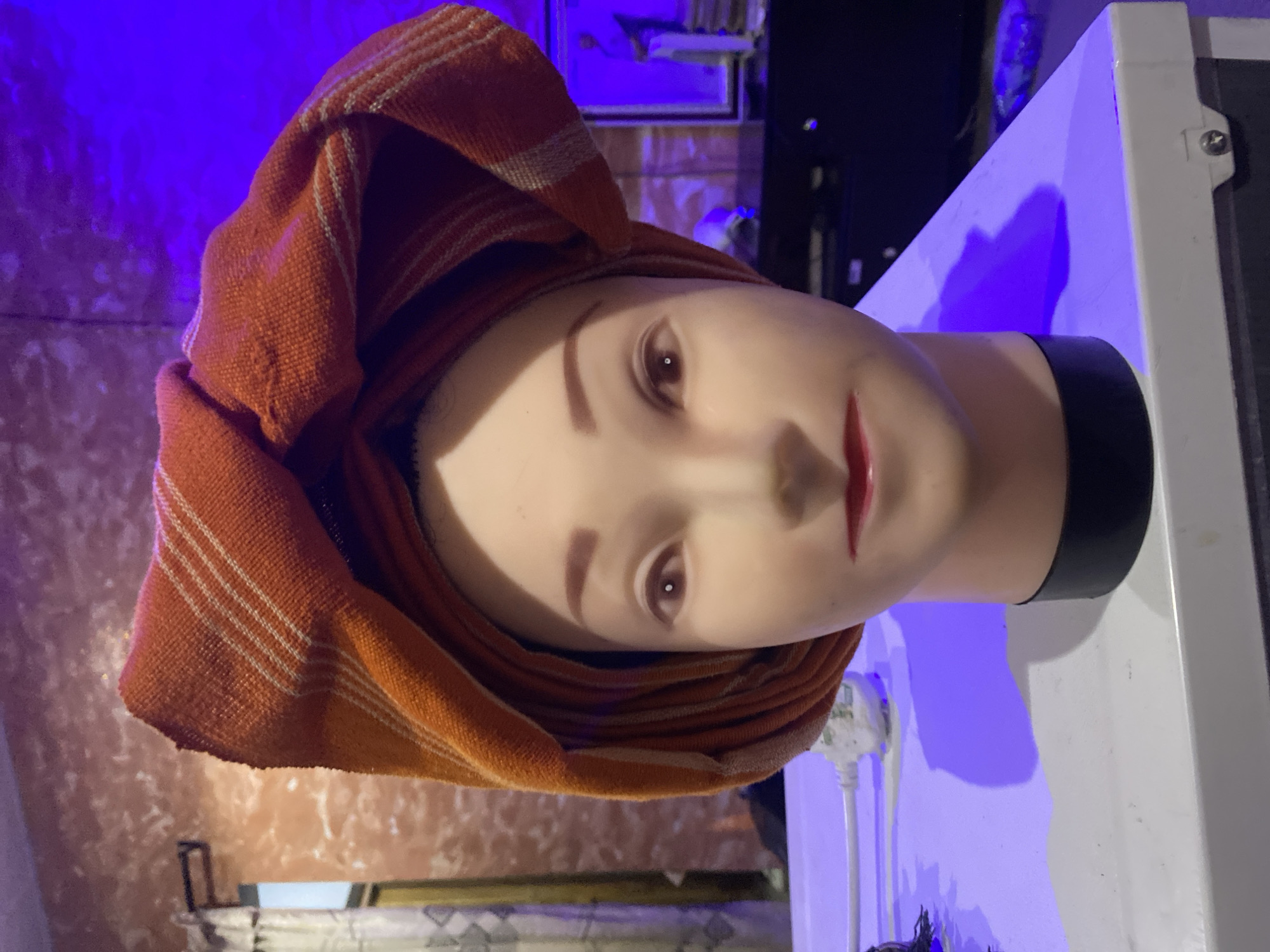 A mannequin head draped in a striped orange towel is the central subject. The mannequin has pale skin, painted eyebrows, brown eyes, and red lips. The base of its neck is black. It sits on a white surface, possibly a table or counter, under a blue light. The background is cluttered, with glimpses of a shelf, artwork, and what appears to be a window. There is no discernible evidence in the image to suggest a location in Badagry, Nigeria.