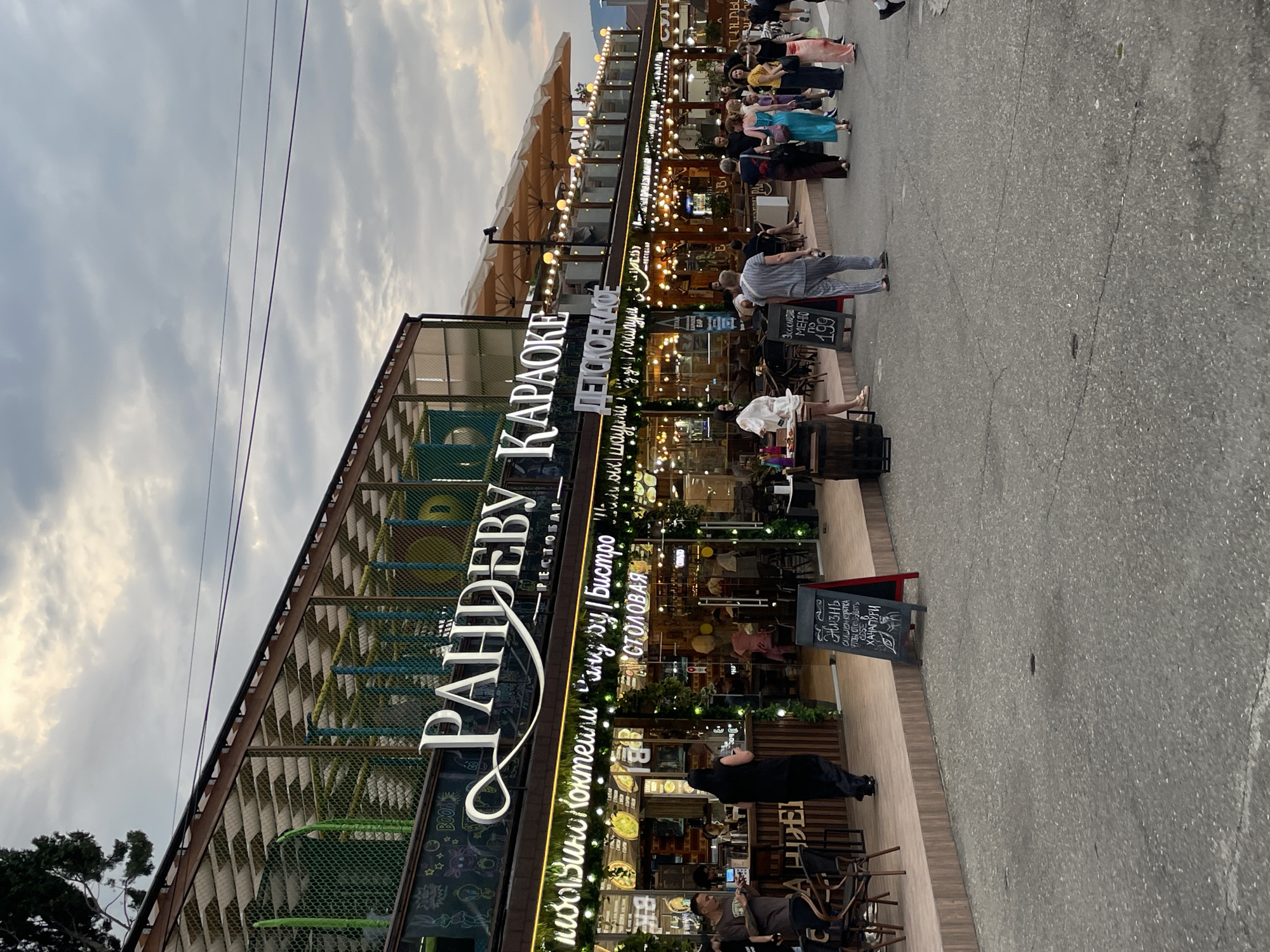 A wide shot shows a street scene outside a multi-story entertainment venue in Sochi, Russia, likely in the evening. The venue has a large sign that reads "PAHDEBY KAPAKOE" and below that, "ДЕТСКОЕ КАФЕ," indicating it is a karaoke bar and a children's cafe. The building has a modern, open-air design with multiple levels, featuring an enclosed play area on the upper floor visible through a wire mesh. The lower level has a prominent facade with string lights, decorated with greenery, and outdoor seating areas. Several people are visible walking and standing on the sidewalk and street in front of the venue. The atmosphere appears casual and social, with individuals dressed in everyday clothing. The ground is asphalt, and the sky is overcast. The overall impression is that of a lively evening establishment in a public area.