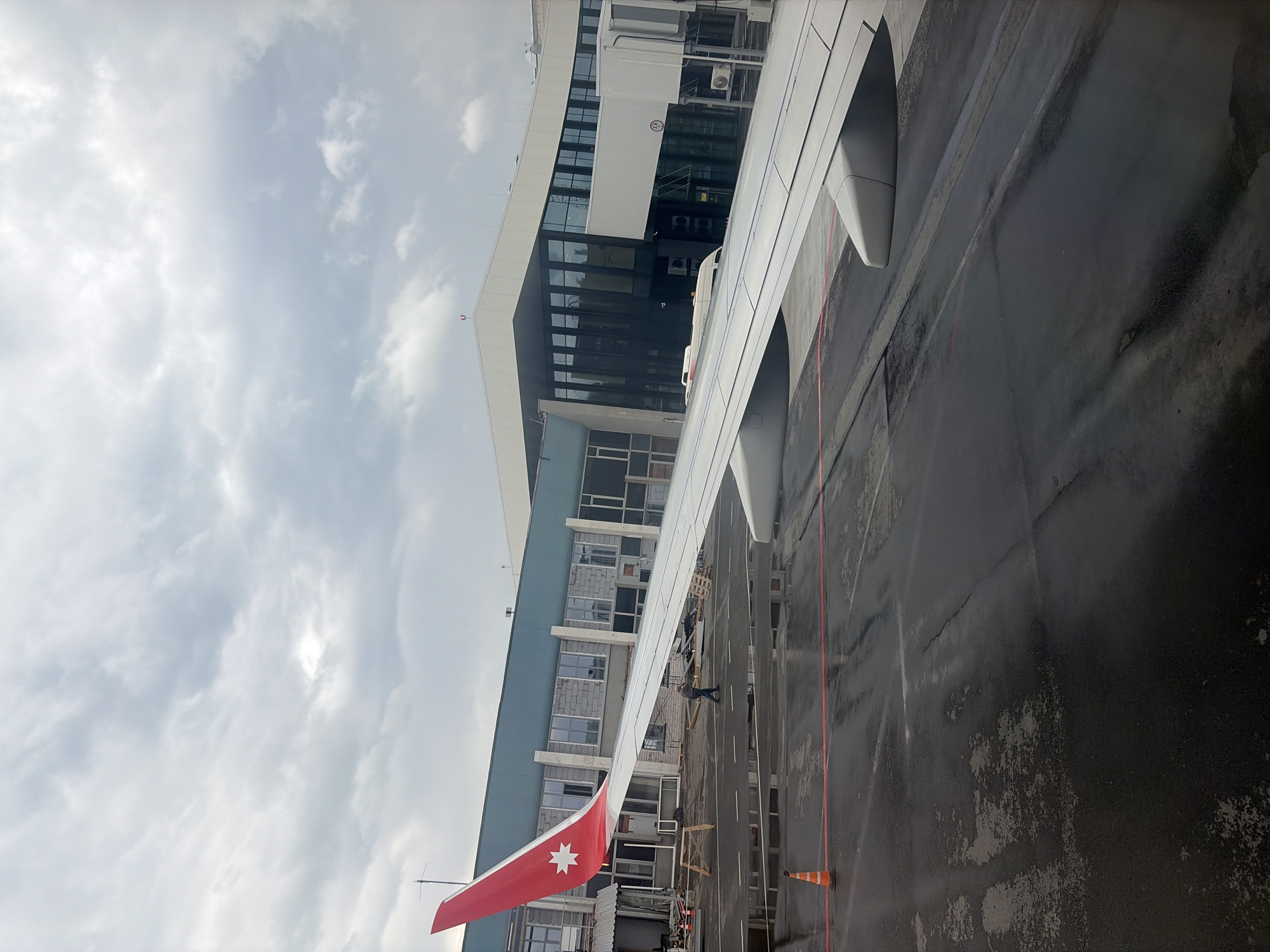 An airplane wing, featuring a distinctive red winglet with a white seven-pointed star (Royal Jordanian Airlines livery), is prominently visible in the foreground, parked on a wet tarmac next to a modern airport terminal building. The view is from inside the aircraft, looking outwards.
The multi-story terminal building, with large glass windows and a contemporary design, dominates the midground. A jet bridge extends from the terminal towards the aircraft, suggesting it is either boarding, disembarking, or preparing for departure/arrival. Several air conditioning units are visible on the exterior of the building.
The tarmac is dark and appears wet, reflecting the overcast, cloudy sky above. White and red lines are painted on the ground, indicating taxiways or parking positions. An orange traffic cone is visible on the tarmac. In one crop, a person appears to be standing near the winglet, and what looks like scaffolding or construction materials are present nearby. A small white ground vehicle or bus is also visible near the jet bridge.
Visible text on the terminal windows includes "DUTY FREE," "LATTE," and "TAX FREE." A circular caution symbol is also seen on the jet bridge. The sky is uniformly gray and cloudy, indicating overcast weather conditions. The scene is set at an airport, consistent with the location context of Новое Мартьяново, Russia.