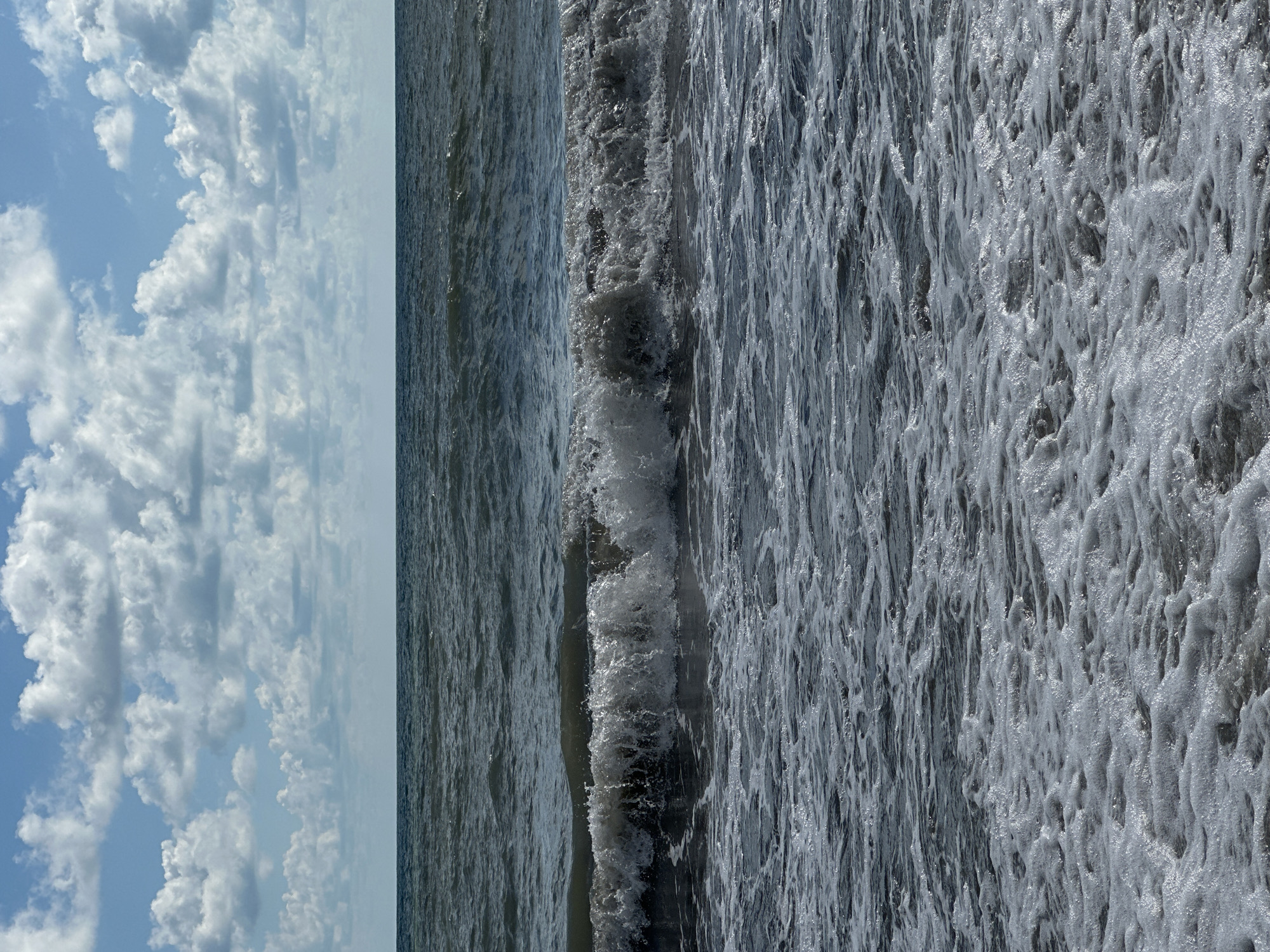 The image depicts a view of the Black Sea from the shore in Lermontovo, Russia. The sky is partly cloudy with white cumulus clouds against a blue sky. The sea is dark and choppy, with a wave breaking in the mid-ground. The foreground is filled with white foam from the receding wave. The horizon line is clearly visible, separating the sea and sky. There are no people or man-made objects visible in the image.