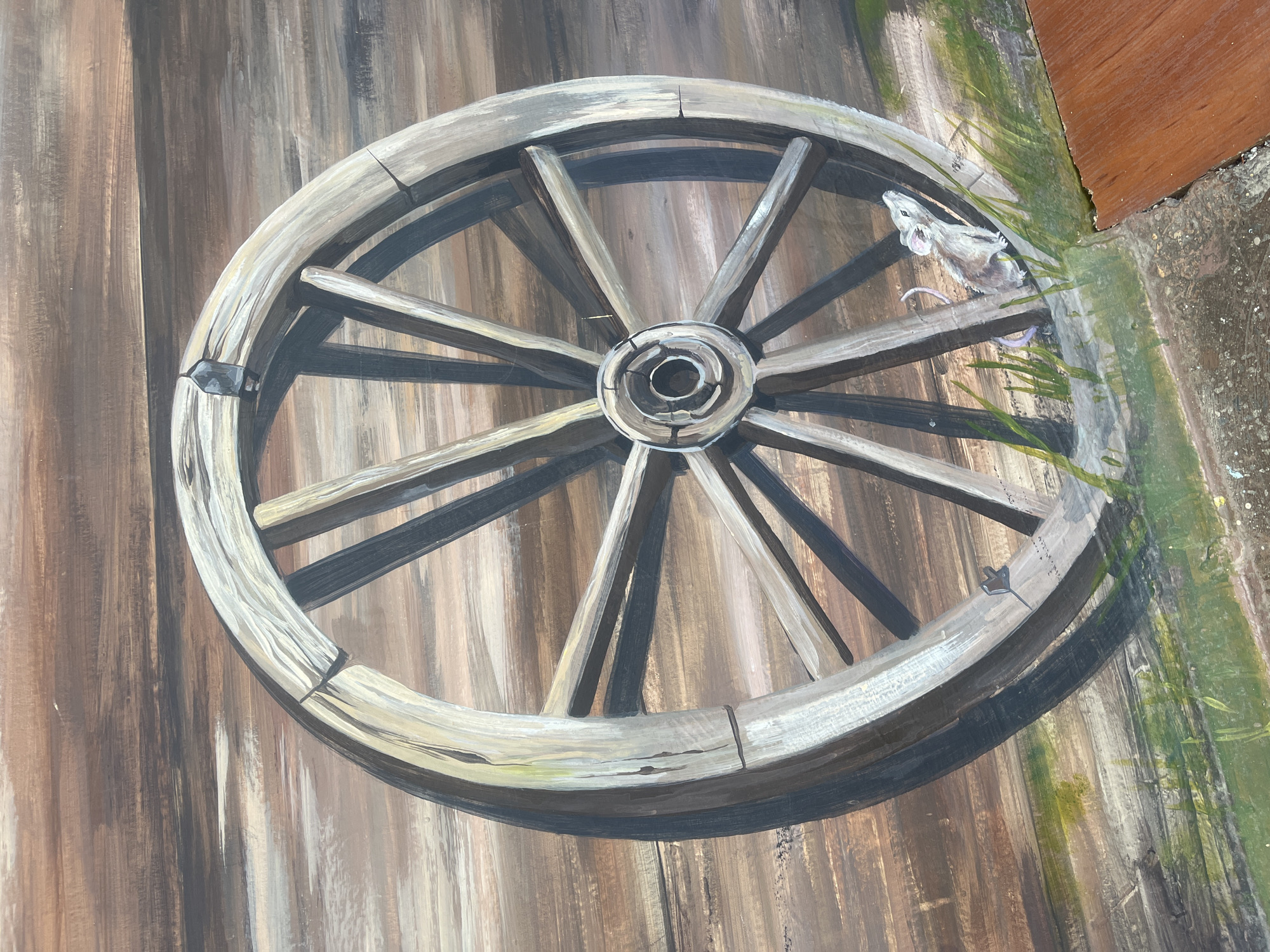 The image displays a painting of a weathered wooden wagon wheel leaning against a wooden wall. The wheel is large and appears to be old, with visible cracks and spokes. A small mouse is depicted at the base of the wheel, peeking out from behind blades of grass. The background wall is made of horizontal wooden planks painted in shades of brown and gray, suggesting an rustic outdoor setting. The lower portion of the image shows a textured surface, possibly concrete or stone, with a section of reddish-brown wood in the bottom right corner. This artwork is located on a wall in the city of Hrodna, Belarus.